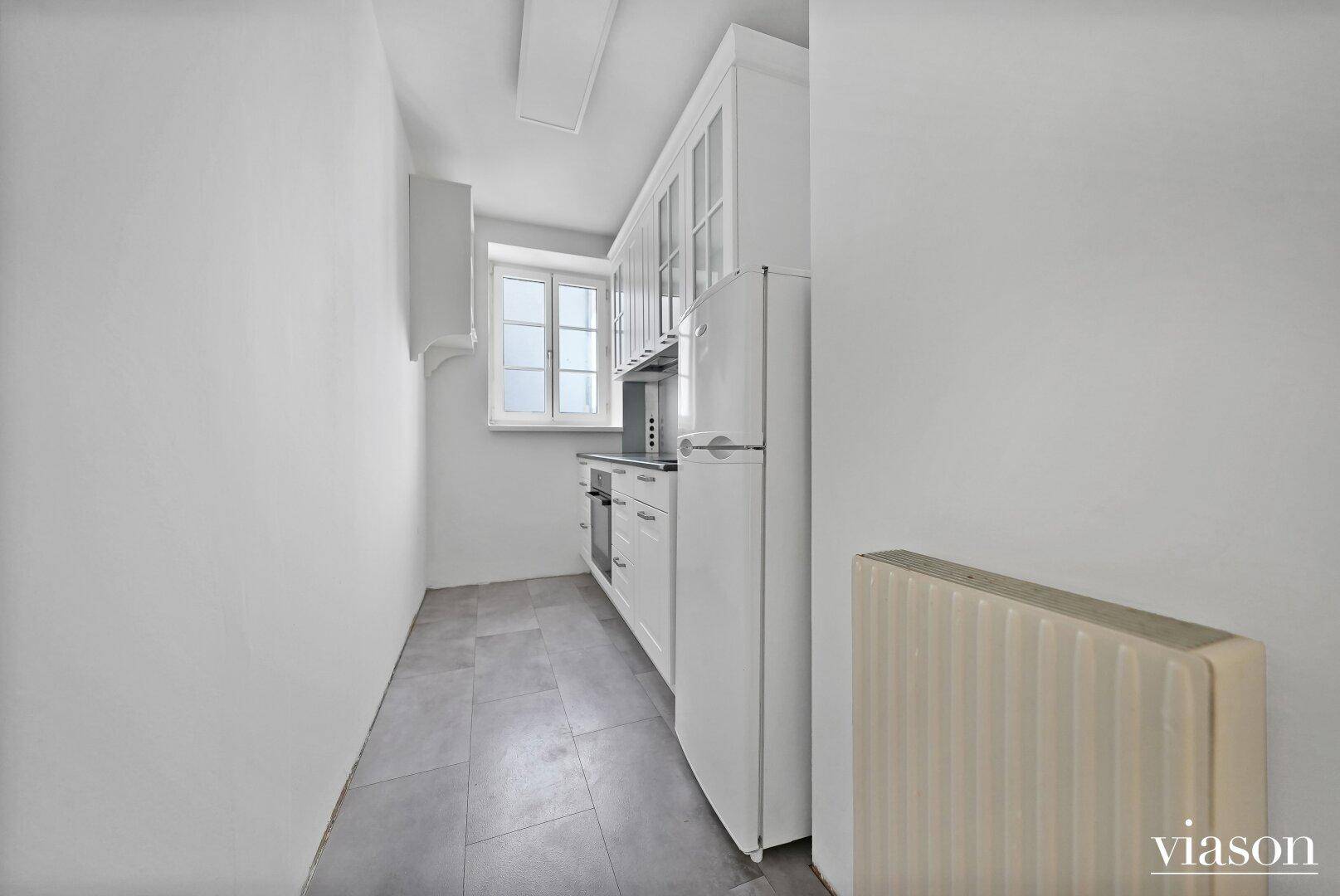 Schmale Küche mit weißen Einbauschränken, Kühlschrank und Fenster, funktional eingerichtet.