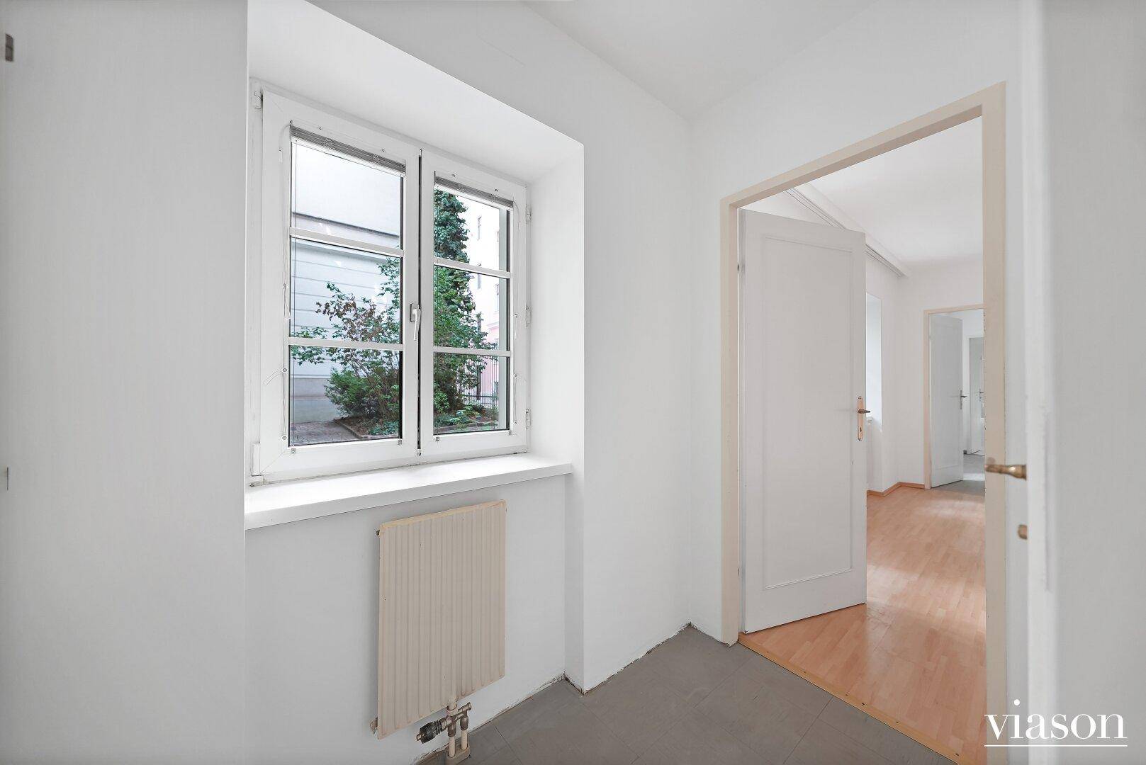 Heller Raum mit Fenster und Heizkörper, der zu einem weiteren Zimmer führt.