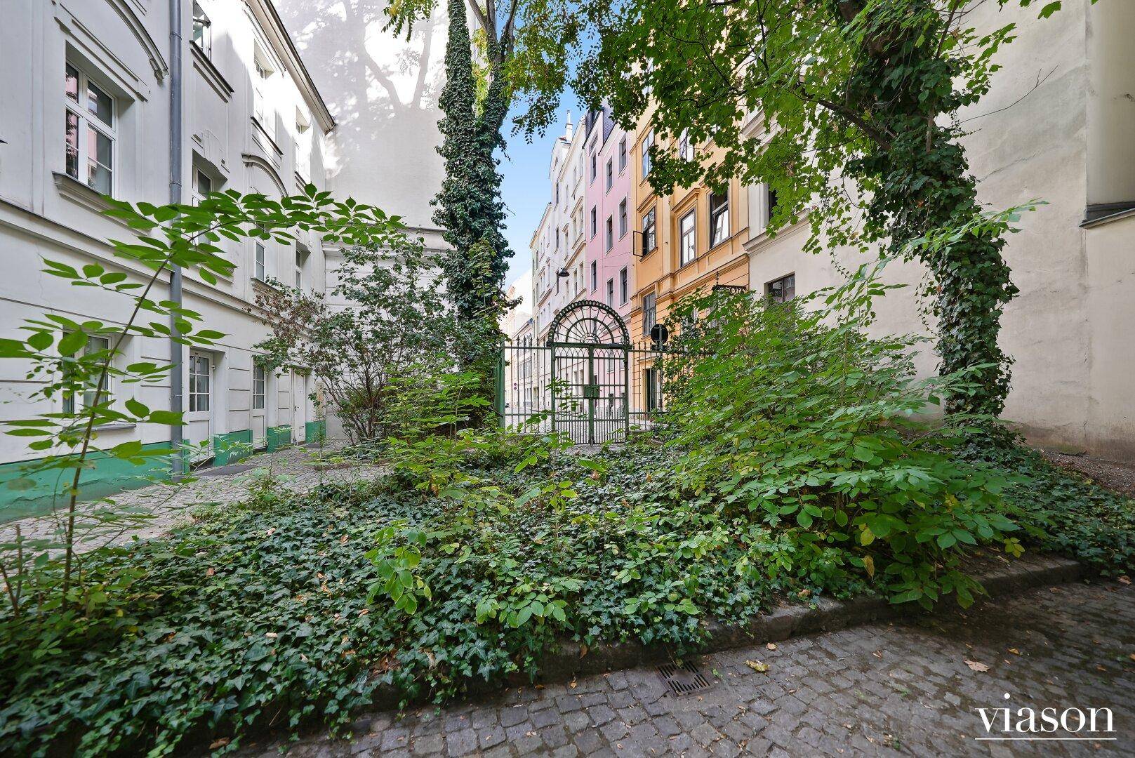 Grüner Innenhof mit üppiger Bepflanzung, Kopfsteinpflaster und historischen Gebäudefassaden.
