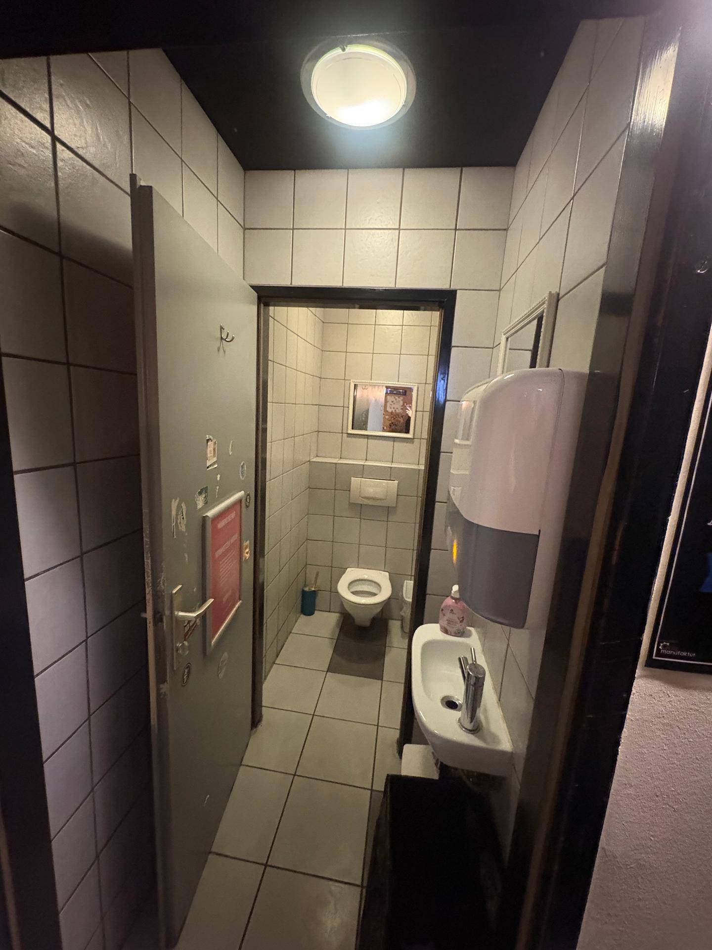 Saubere öffentliche Toilette mit weißen Fliesen, Wand-WC und kleinem Waschbecken.