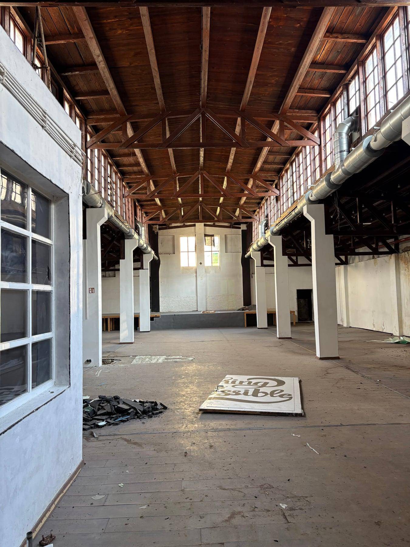 Große, leere Halle im Industriestil mit hohen Holzbalkendecken, vielen Fenstern und Betonboden.