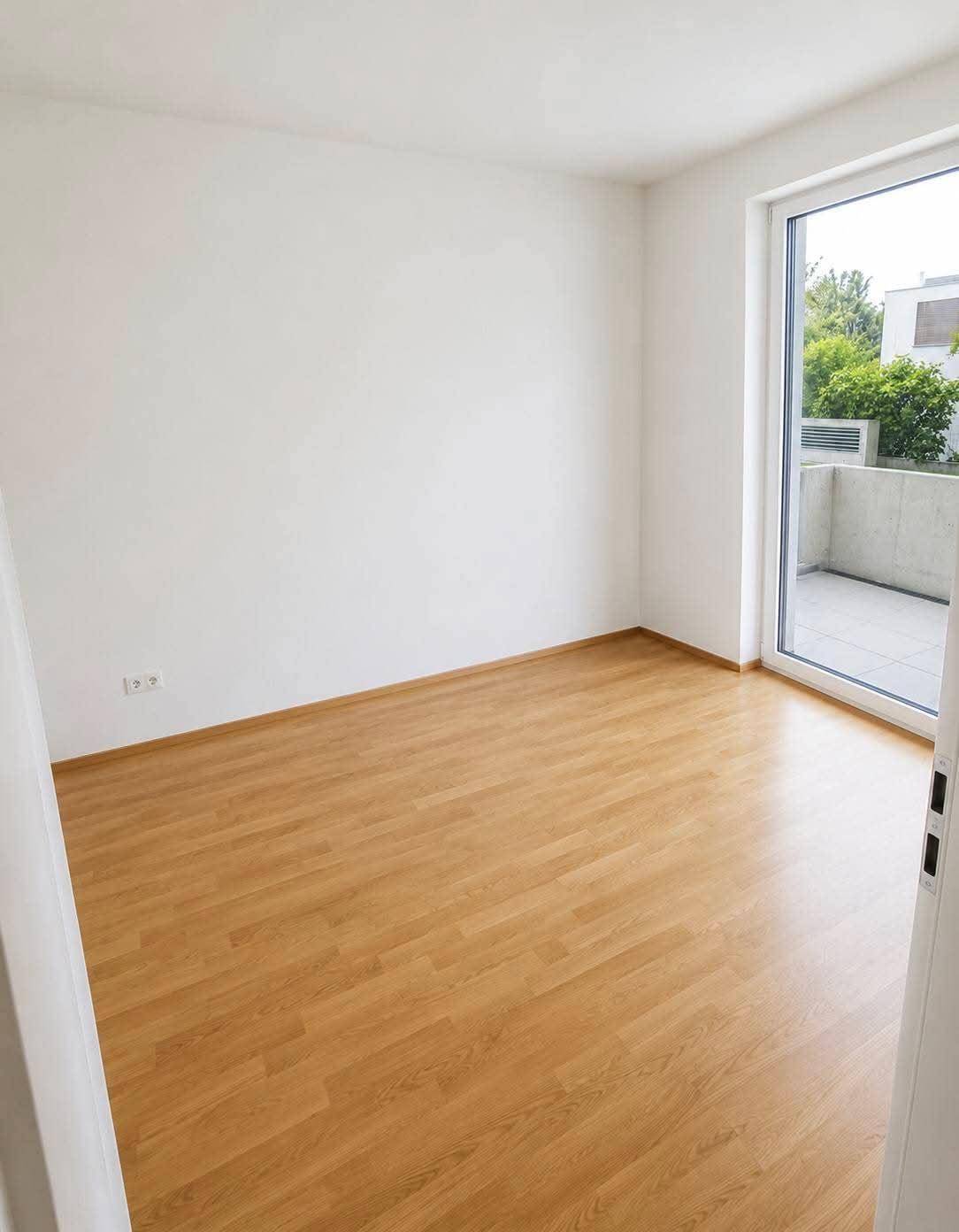 Leeres Schlafzimmer mit hellem Holzboden und großem Fenster mit Zugang zu einem Balkon.