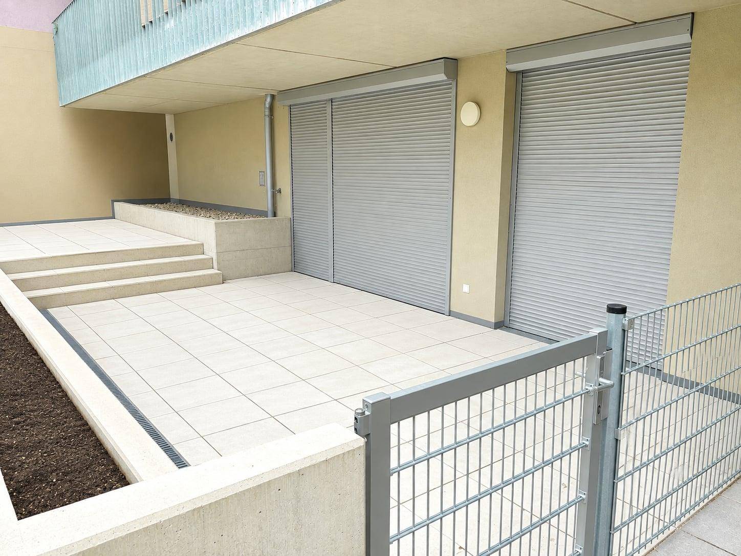 Geräumige geflieste Terrasse mit Stufen, erhöhten Pflanzbeeten und Rollläden an der Fassade.
