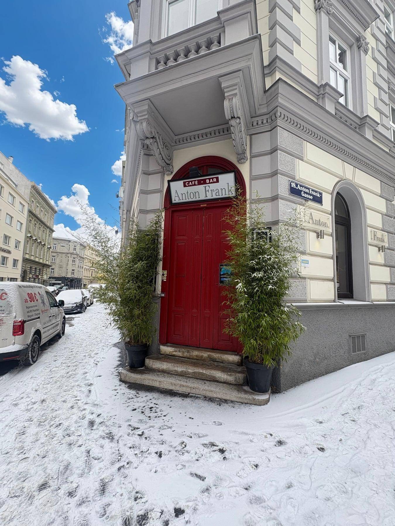 Historische Fassade eines Gebäudes mit roter Eingangstür und Schnee auf dem Bürgersteig unter blauem Himmel.