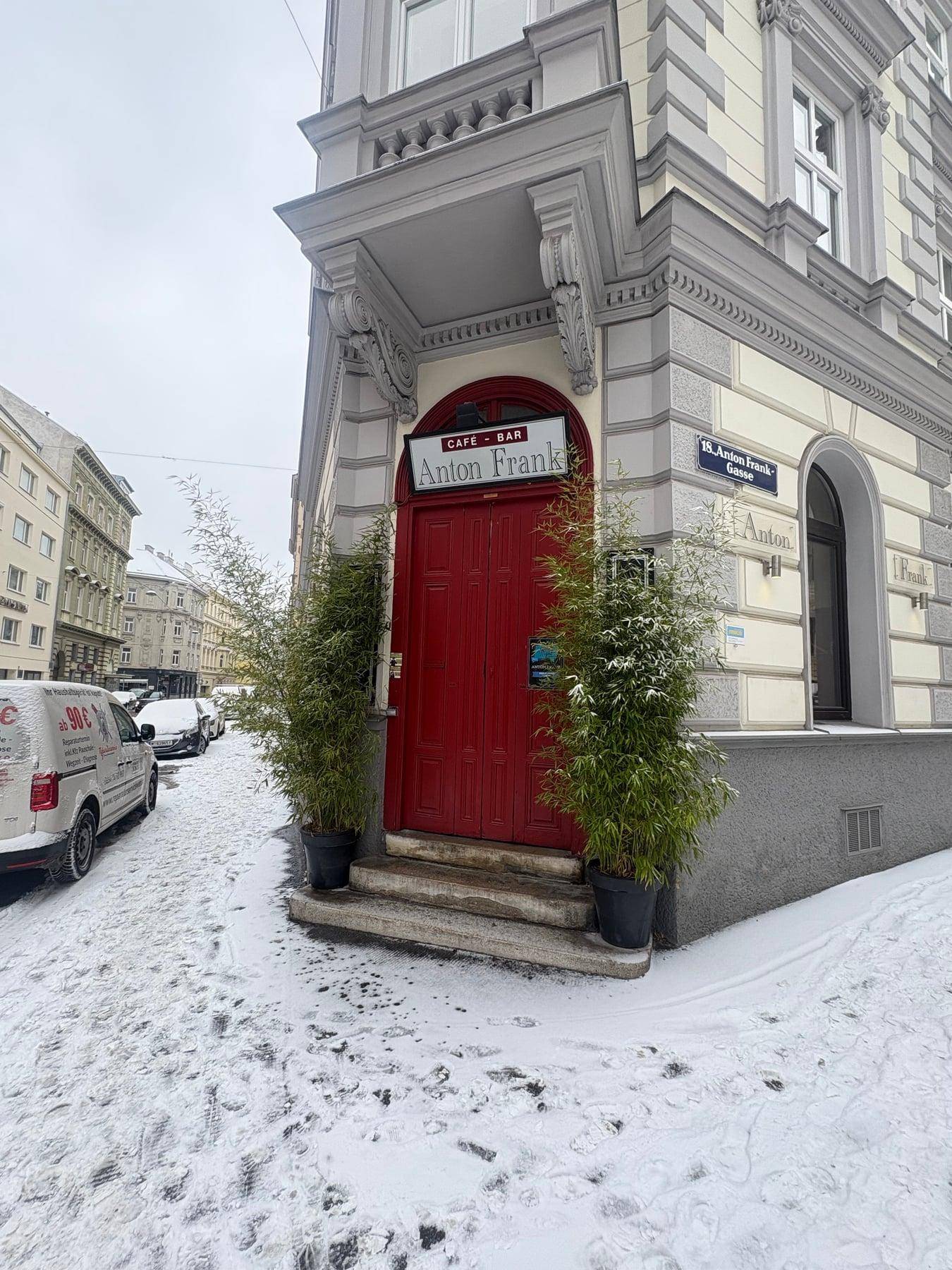 Eingangsbereich eines Gebäudes mit roter Tür, Pflanzen und Schnee auf dem Bürgersteig.