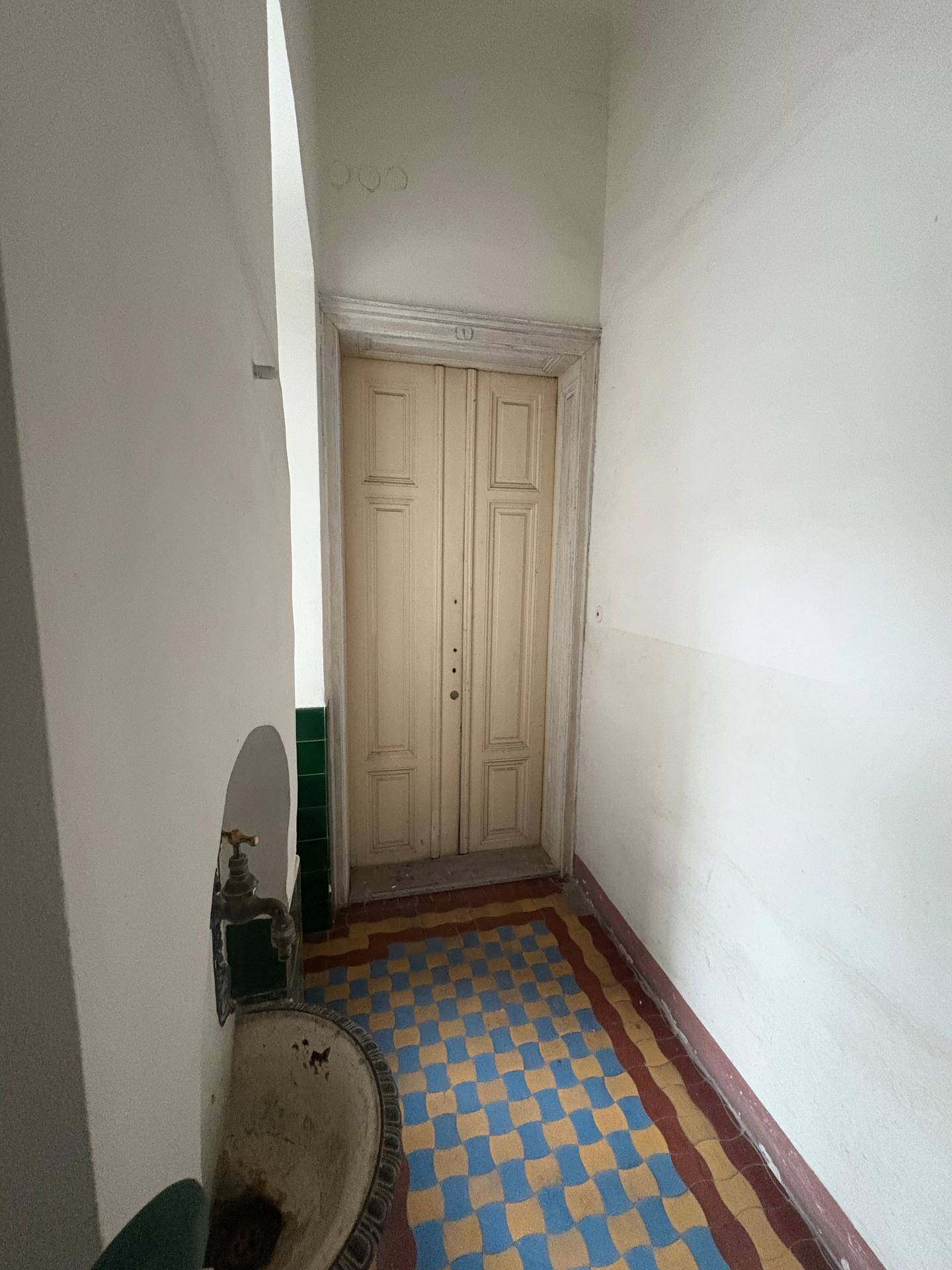 Schmaler Flur in einem Altbau mit einer alten Holztür und einem kleinen Waschbecken.