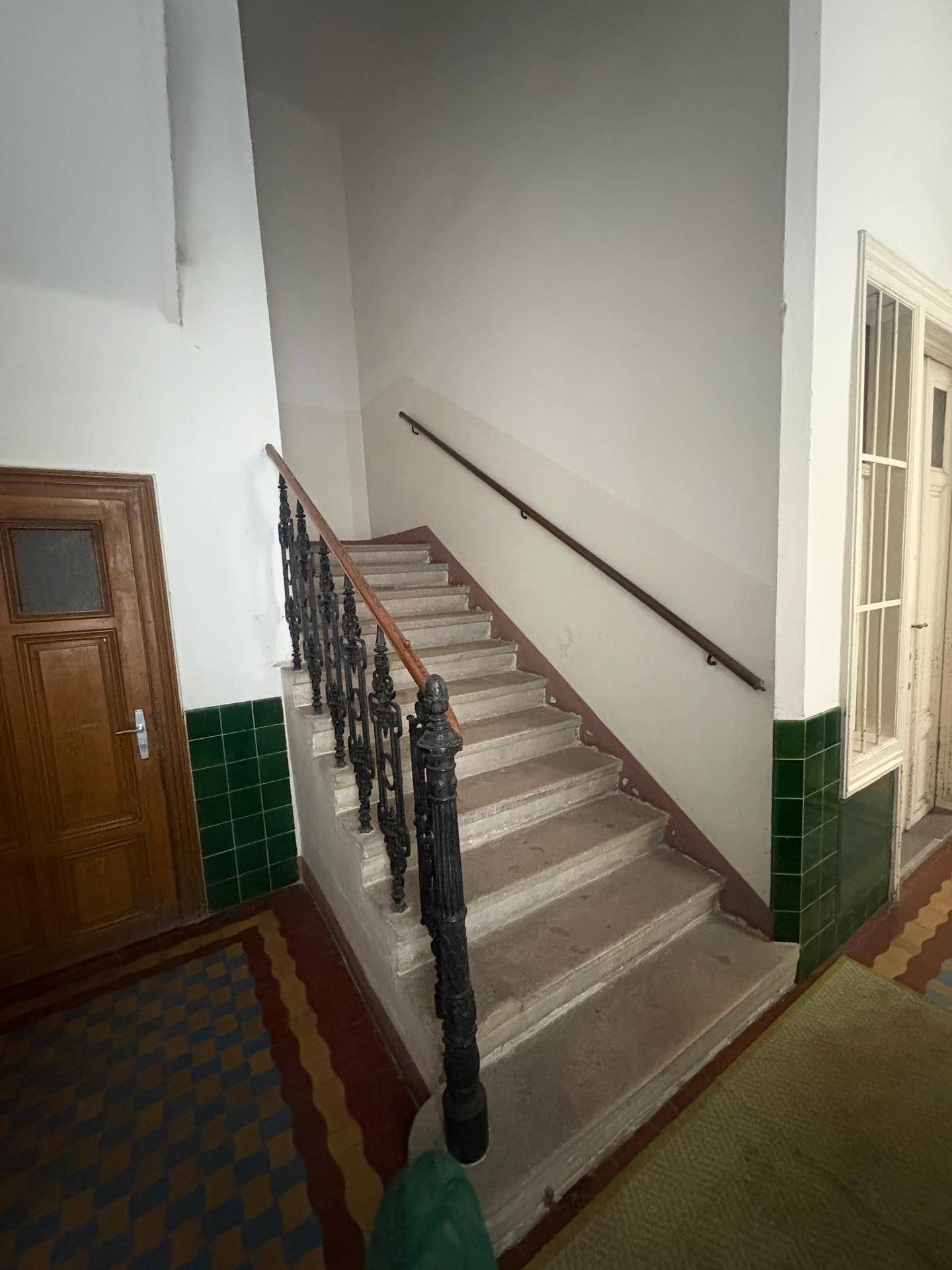 Treppenhaus mit Steinstufen, Holzgeländer und dekorativem schmiedeeisernem Element in einem Altbau.