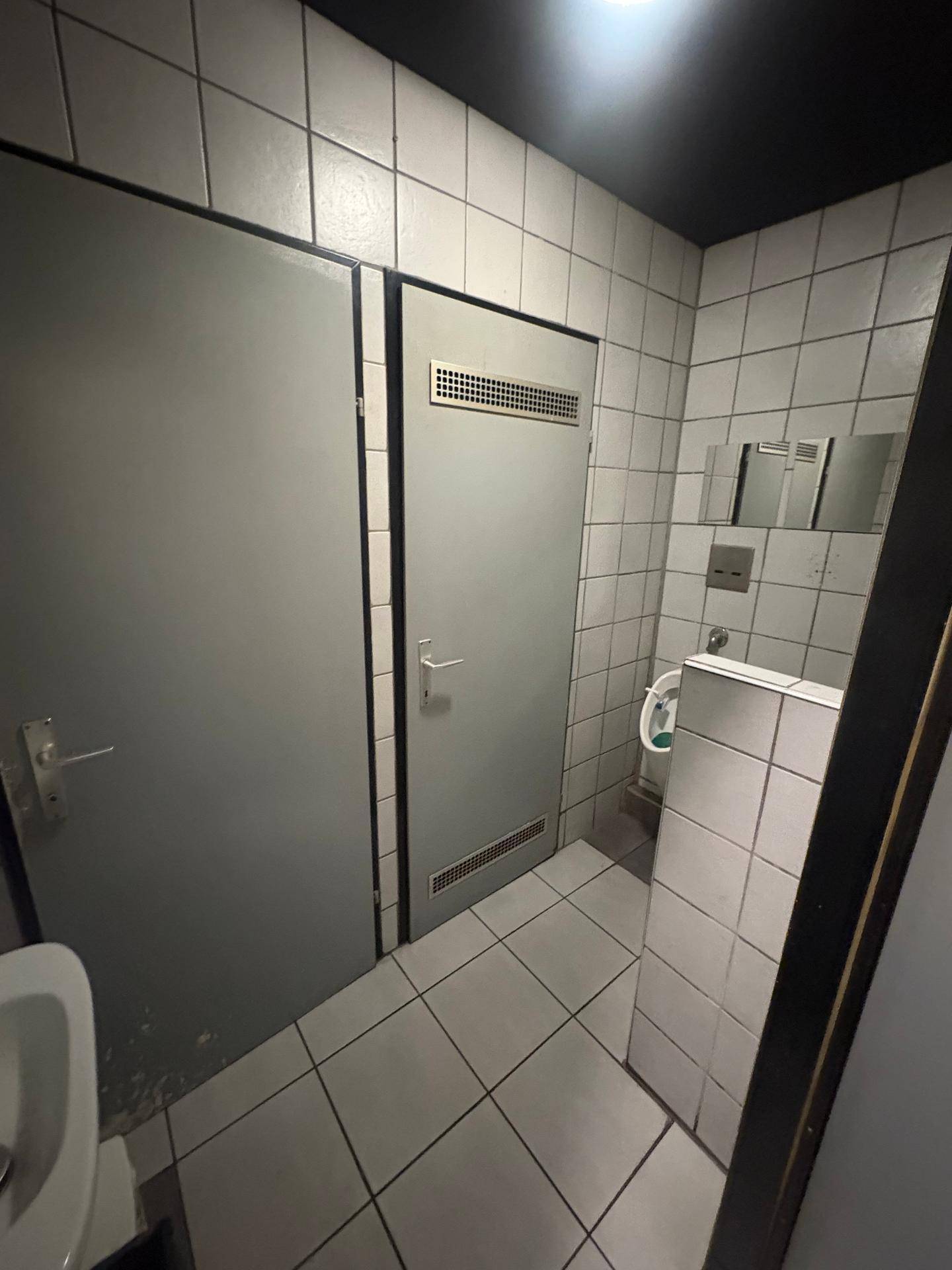 Öffentlicher Toilettenbereich mit grauen Türen, weißen Fliesen und einem Spiegel.