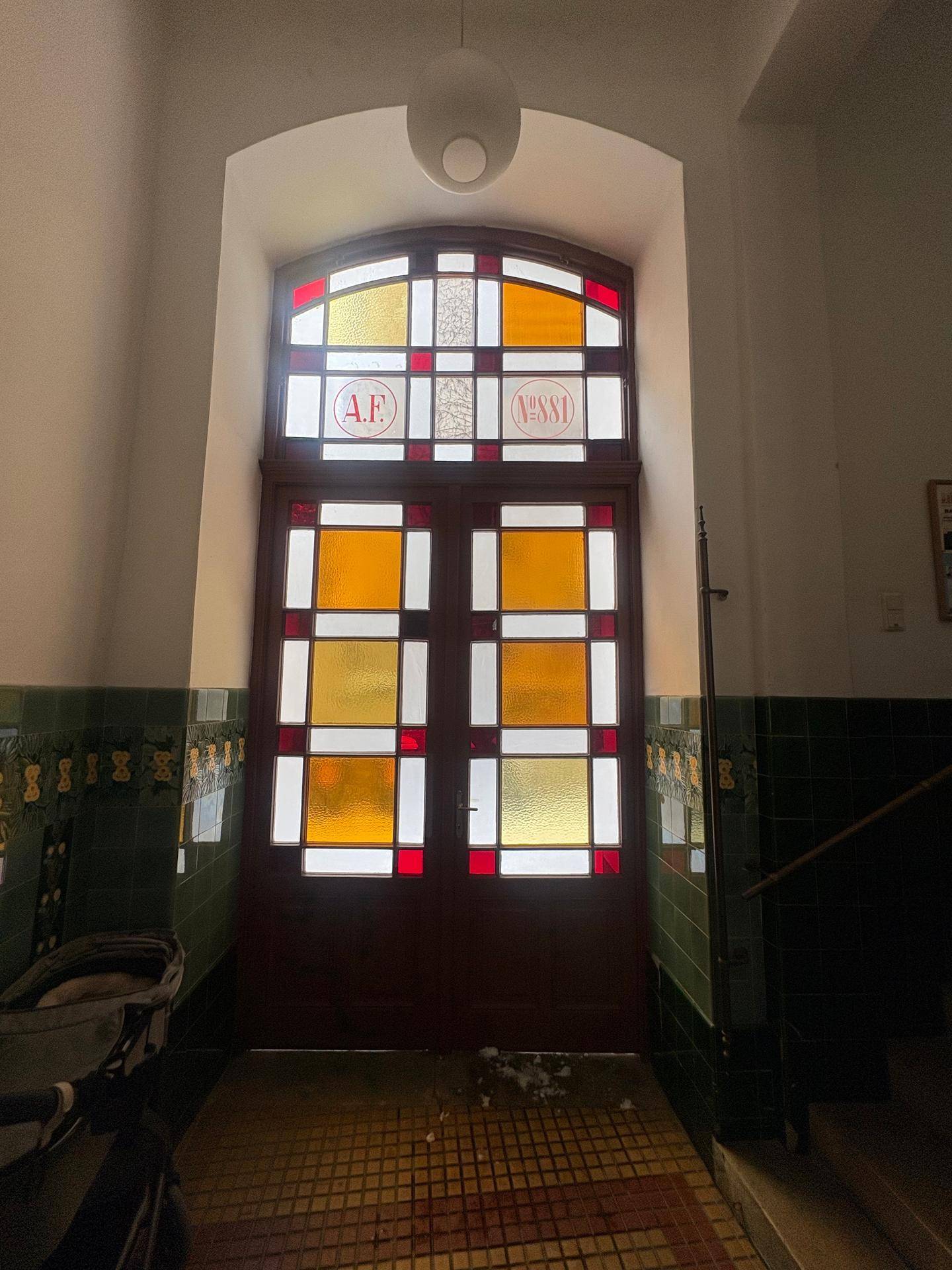 Eingangsbereich mit einer großen Tür aus Buntglas und dekorativen grünen Wandfliesen in einem Altbau.