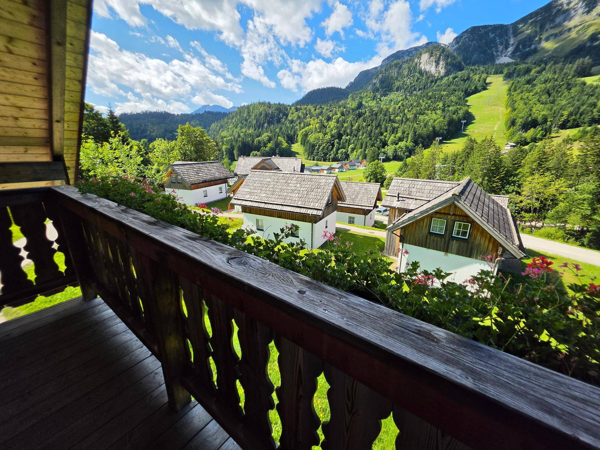 Balkon mit Holzboden und Geländer, bietet einen weiten Blick auf die umliegende Berglandschaft und Häuser.