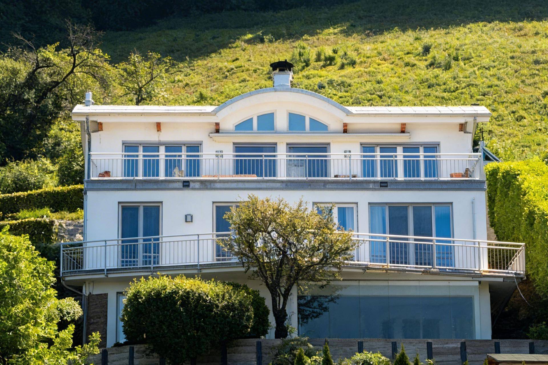 Elegante weiße Villa mit Balkonen und großen Glasfronten, eingebettet in grüne Natur.