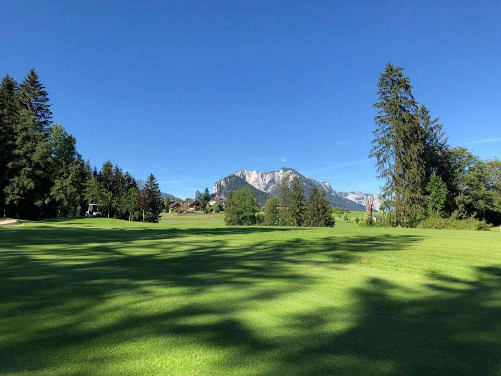 Weitläufige grüne Landschaft mit einem Golfplatz und majestätischen Bergen im Hintergrund unter klarem blauem Himmel.