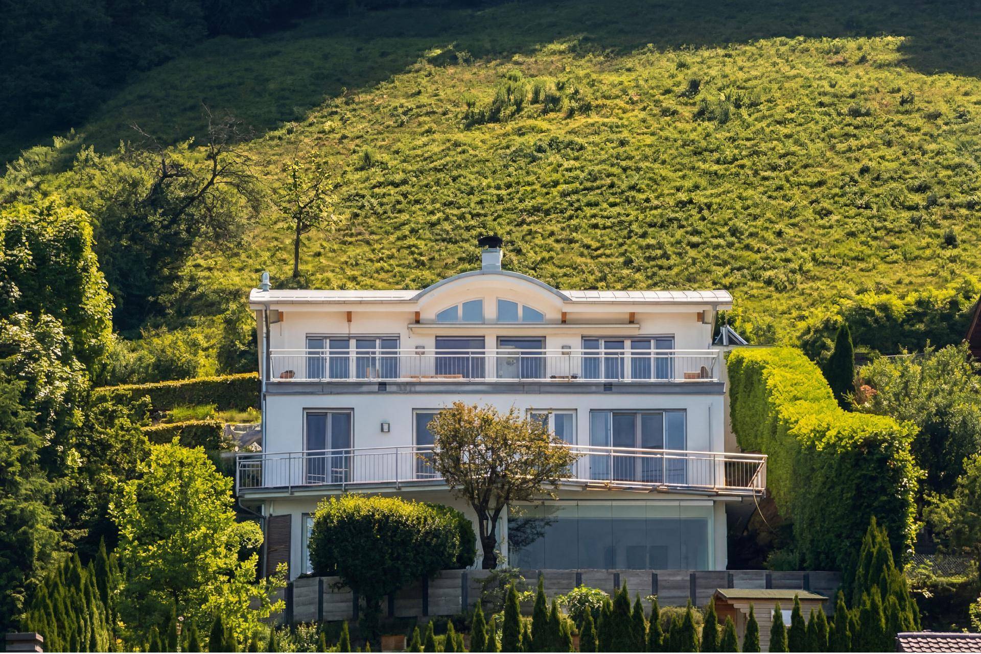 Weiße moderne Villa mit mehreren Balkonen und großen Fenstern, umgeben von grünen Hügeln.