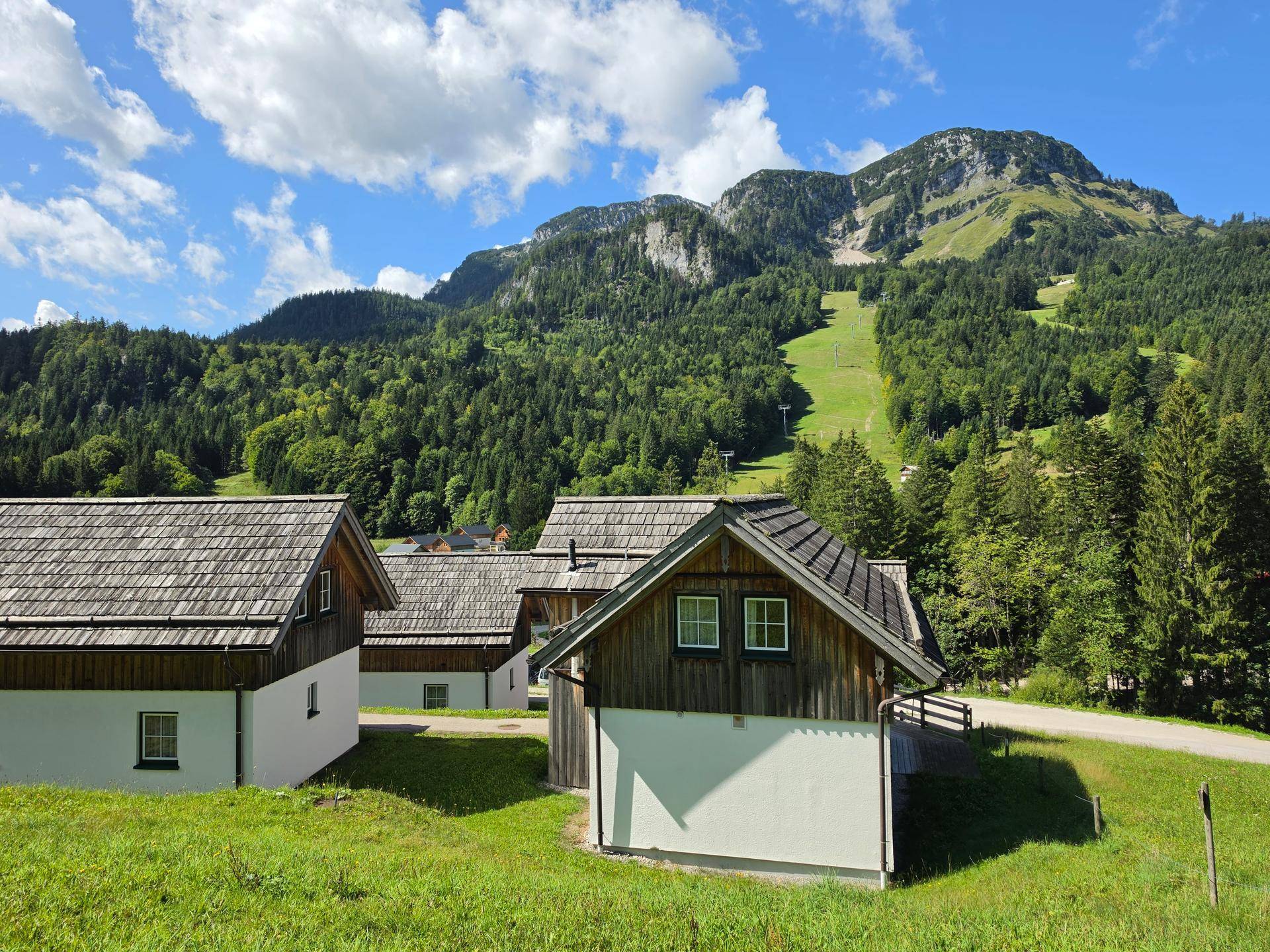 Mehrere traditionelle Holzhäuser in einer grünen Berglandschaft unter blauem Himmel mit Wolken.