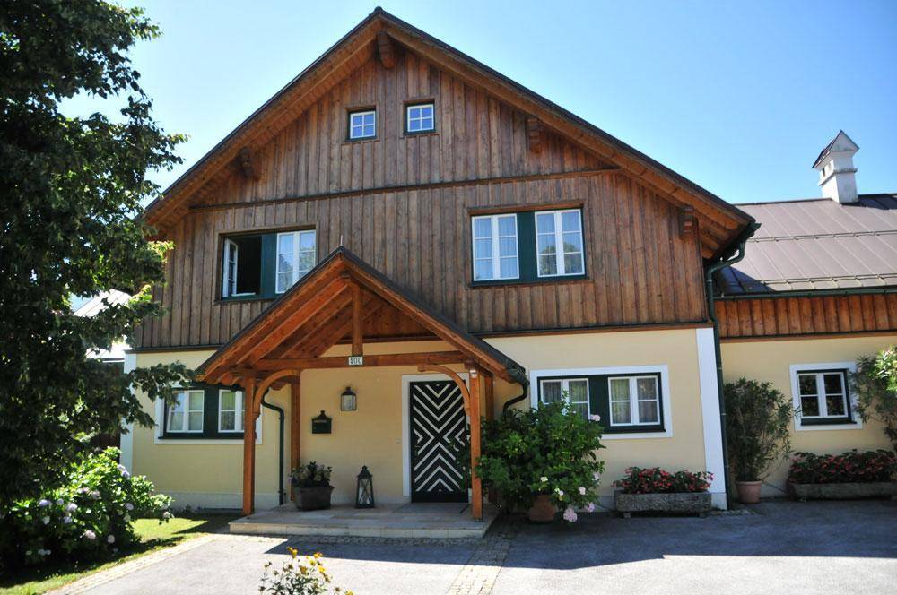 Charmantes Landhaus mit Holzelementen, grünen Fensterläden und einem überdachten Eingangsbereich unter strahlendem Himmel.