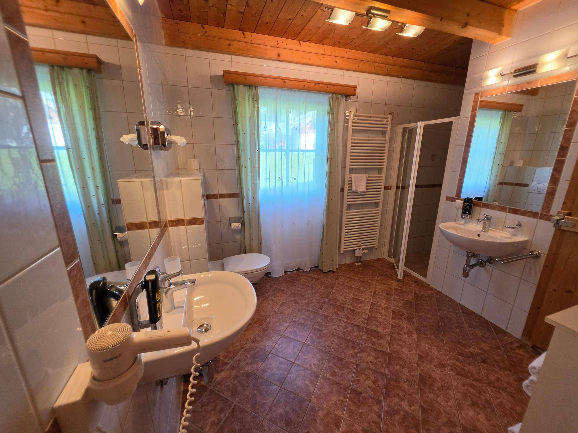 Badezimmer mit braunen Fliesen, zwei Waschbecken, Duschkabine und Fenster, alles in gutem Zustand.