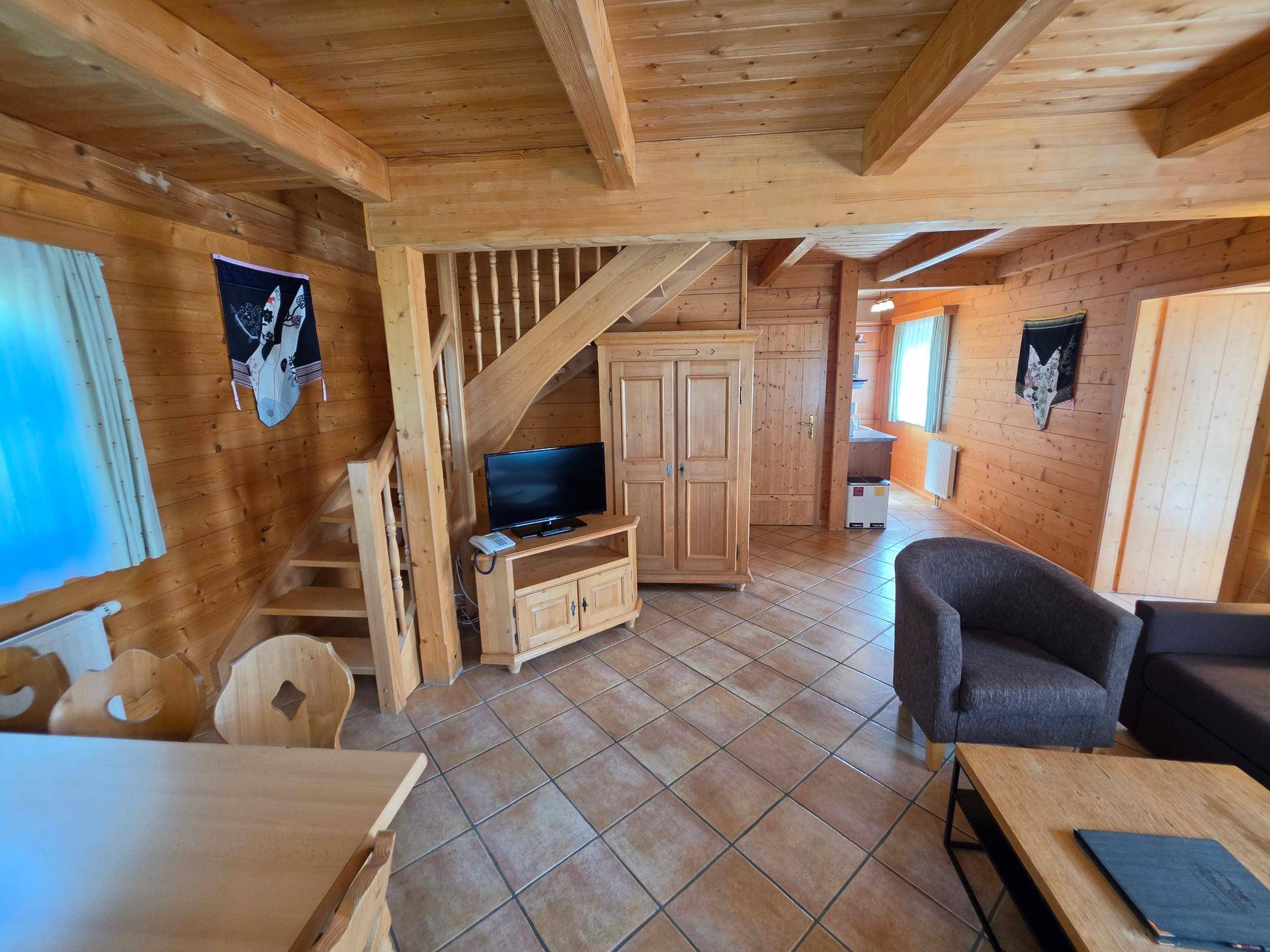 Rustikaler Wohnbereich mit Holzvertäfelung, sichtbaren Balken, gefliestem Boden und Holztreppe zum Obergeschoss.