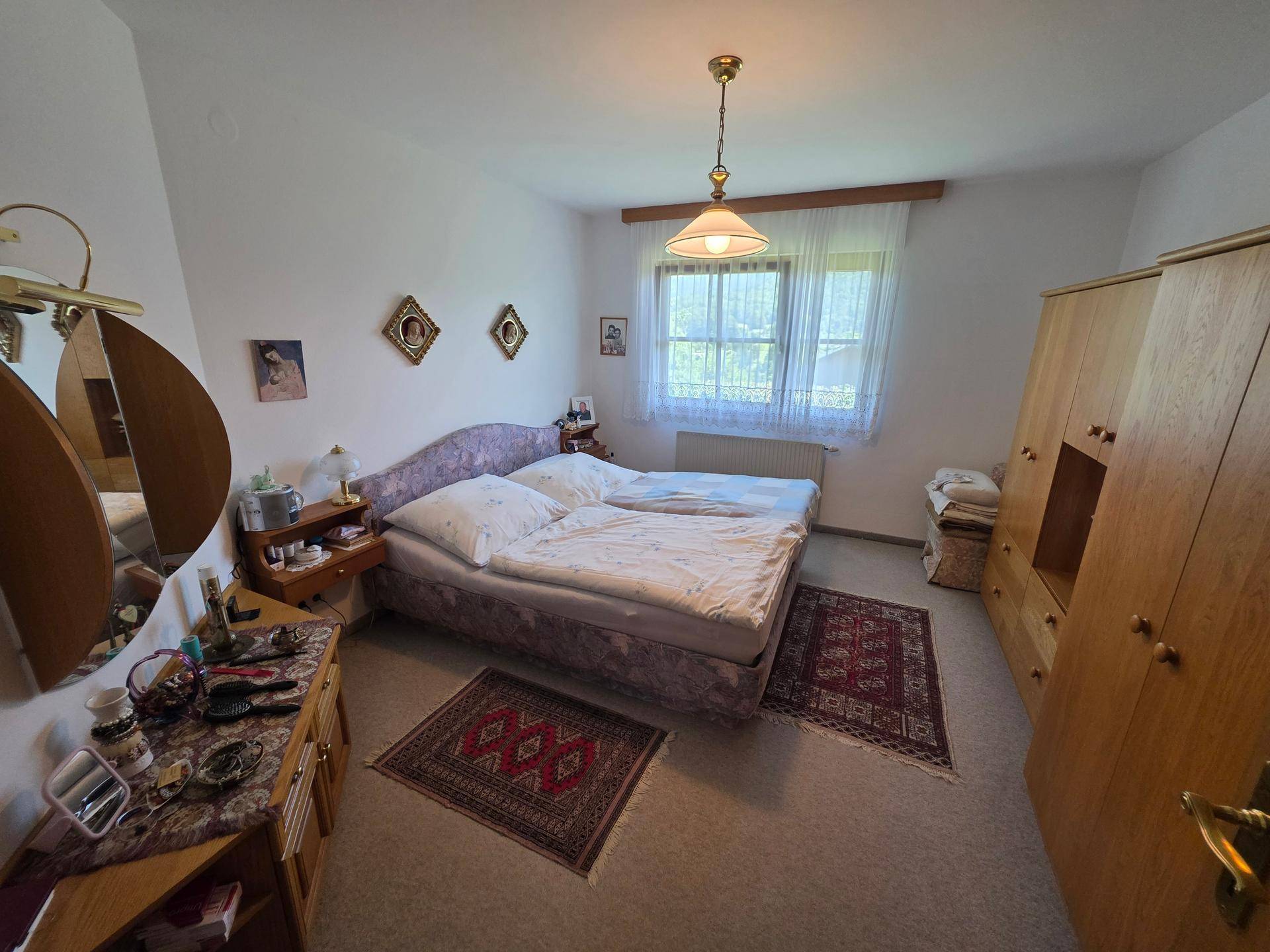 Helles Schlafzimmer mit Doppelbett, Holzschrank und Teppichboden, mit Blick ins Grüne.