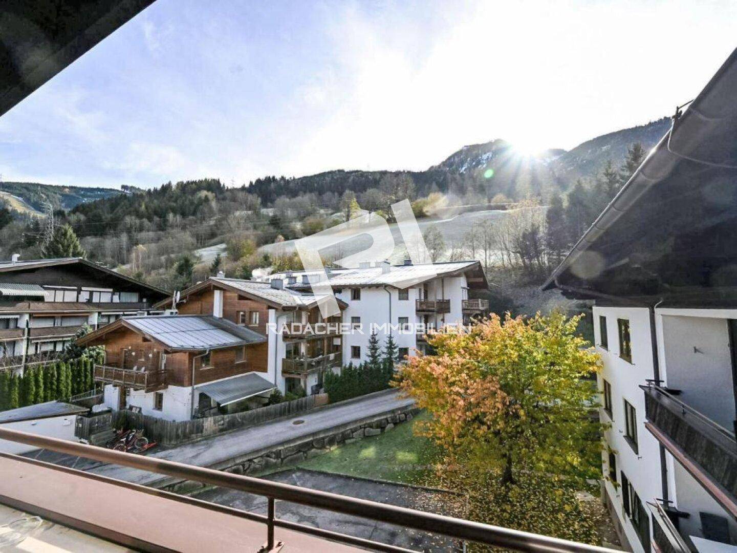 Sonniger Ausblick vom Balkon auf die umliegenden Häuser, Bäume und die majestätische Berglandschaft.