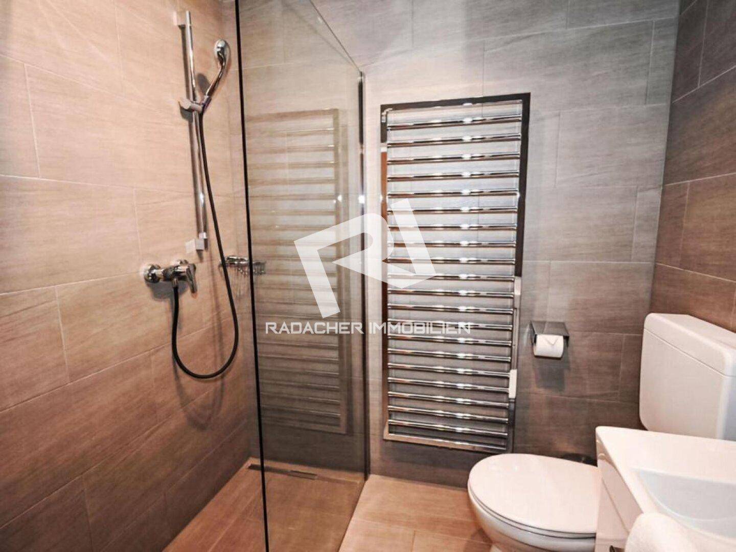 Modernes Badezimmer mit ebenerdiger Dusche, WC und beheiztem Handtuchhalter, alles in Grautönen gehalten.