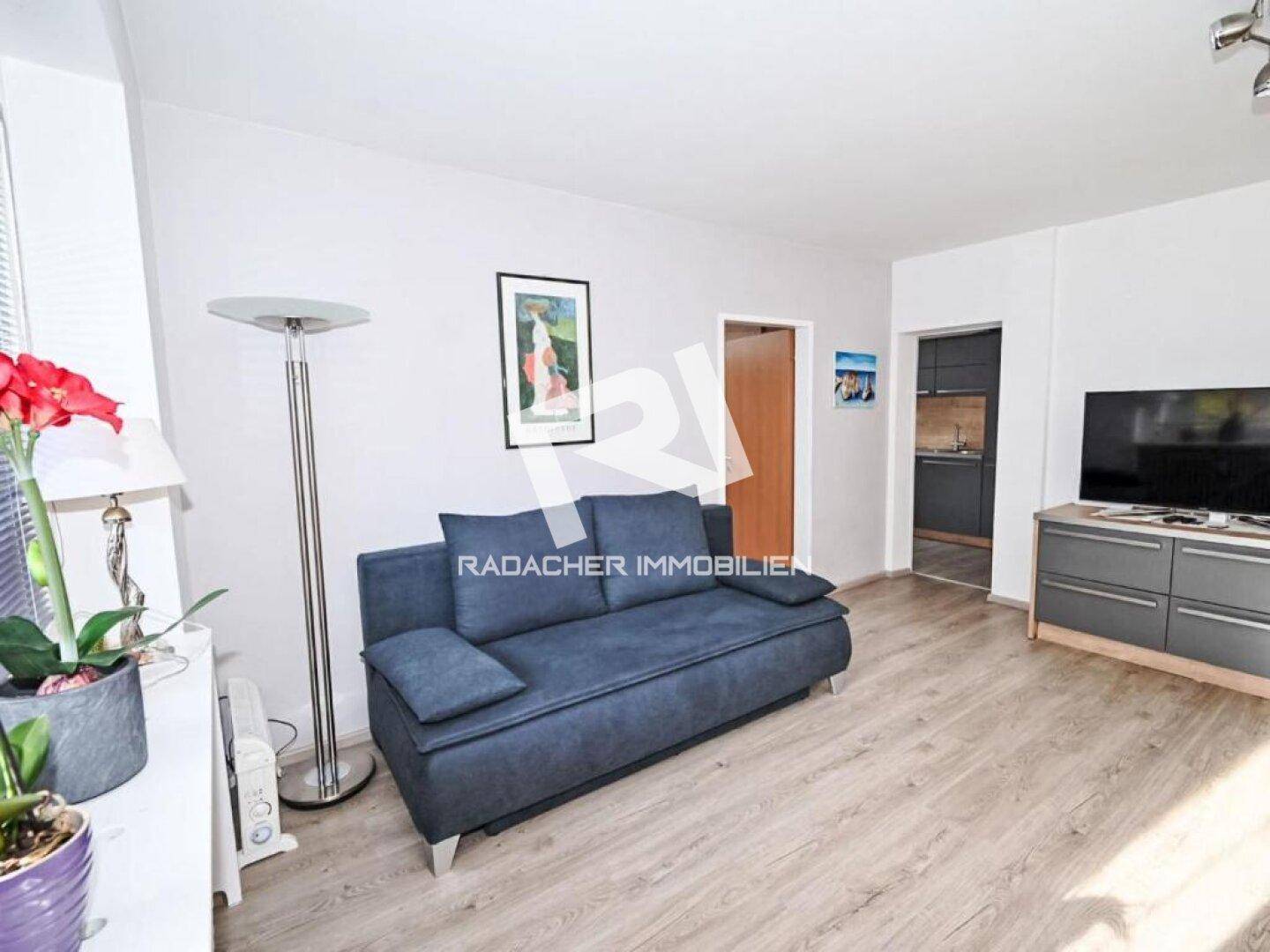 Komfortables Wohnzimmer mit blauem Sofa, Fernseher und Zugang zur Küche, ideal zum Entspannen.