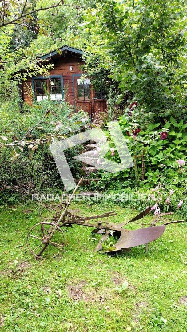 Rustikales Gartenhaus aus Holz, umgeben von üppiger Vegetation und einem alten Pflug.