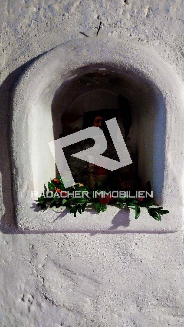 Kleine, weiß verputzte Nische mit religiöser Figur und Blumendekoration in einer Außenwand.