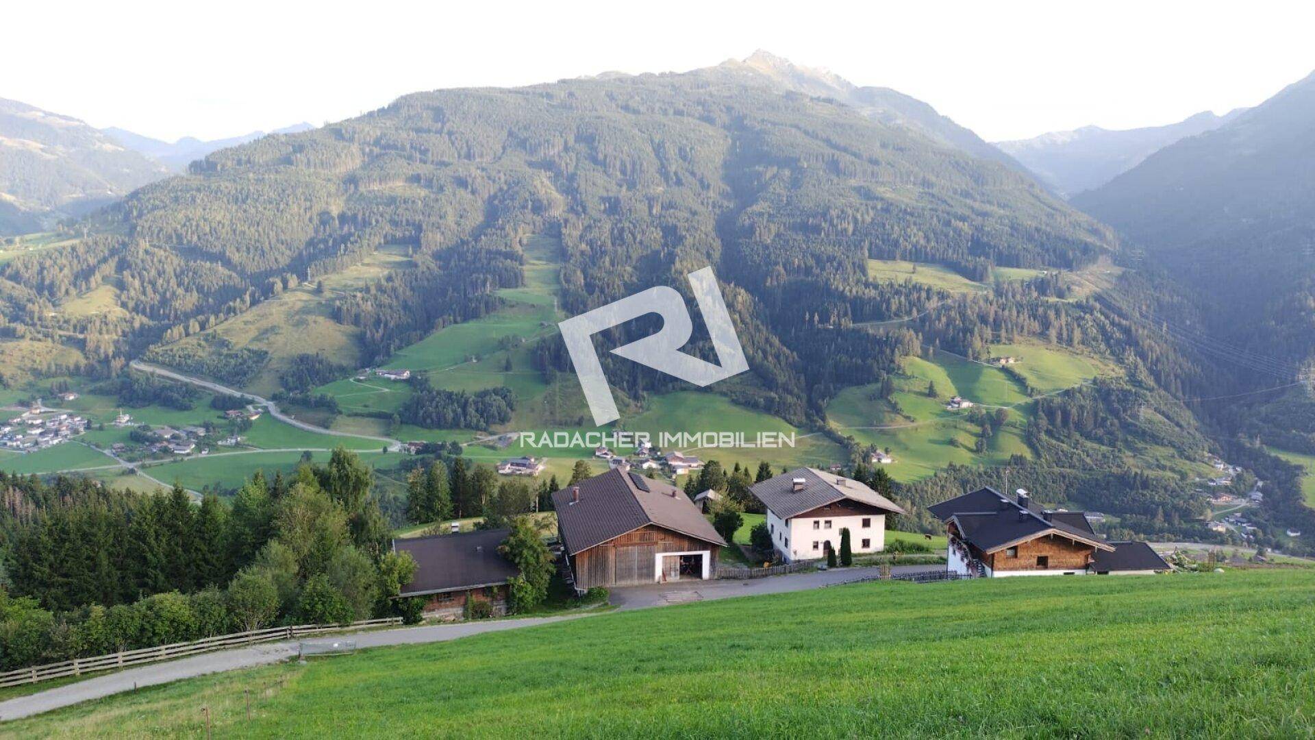 Panoramablick auf eine ländliche Siedlung in einem grünen Tal, umgeben von bewaldeten Bergen.