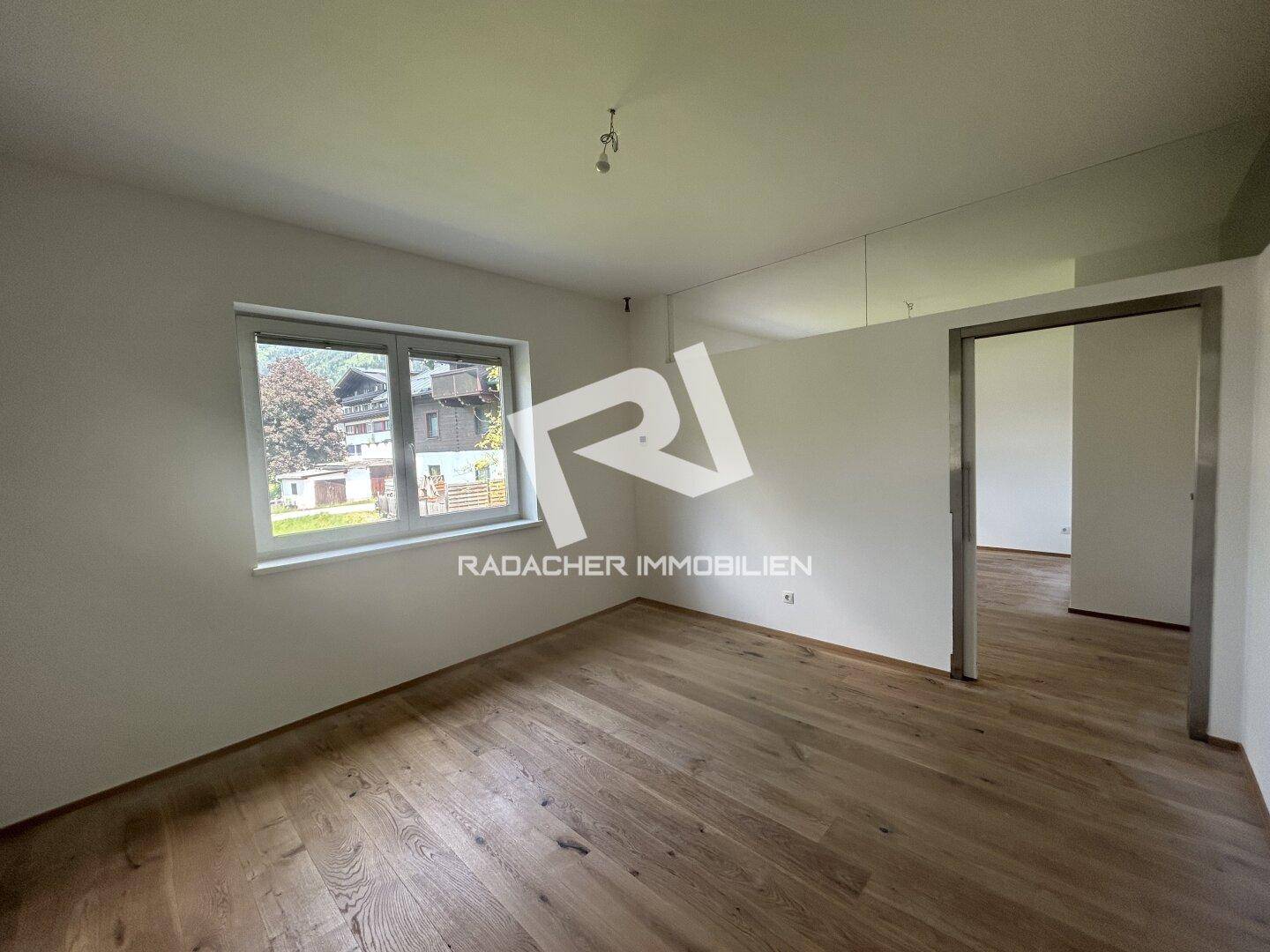Geräumiges, unmöbliertes Zimmer mit Holzboden und Fenster, das einen Blick nach draußen bietet.