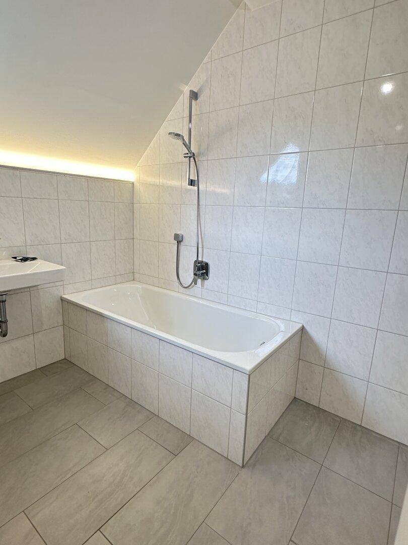 Badezimmer mit Badewanne, Duschfunktion, weißen Wandfliesen und schräger Decke im Dachgeschoss.