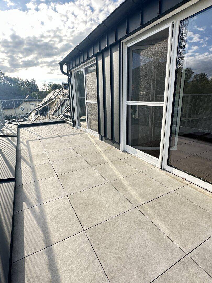 Sonnige Terrasse mit modernen Fliesen, großen Glasfronten und Blick auf den Himmel und die Umgebung.