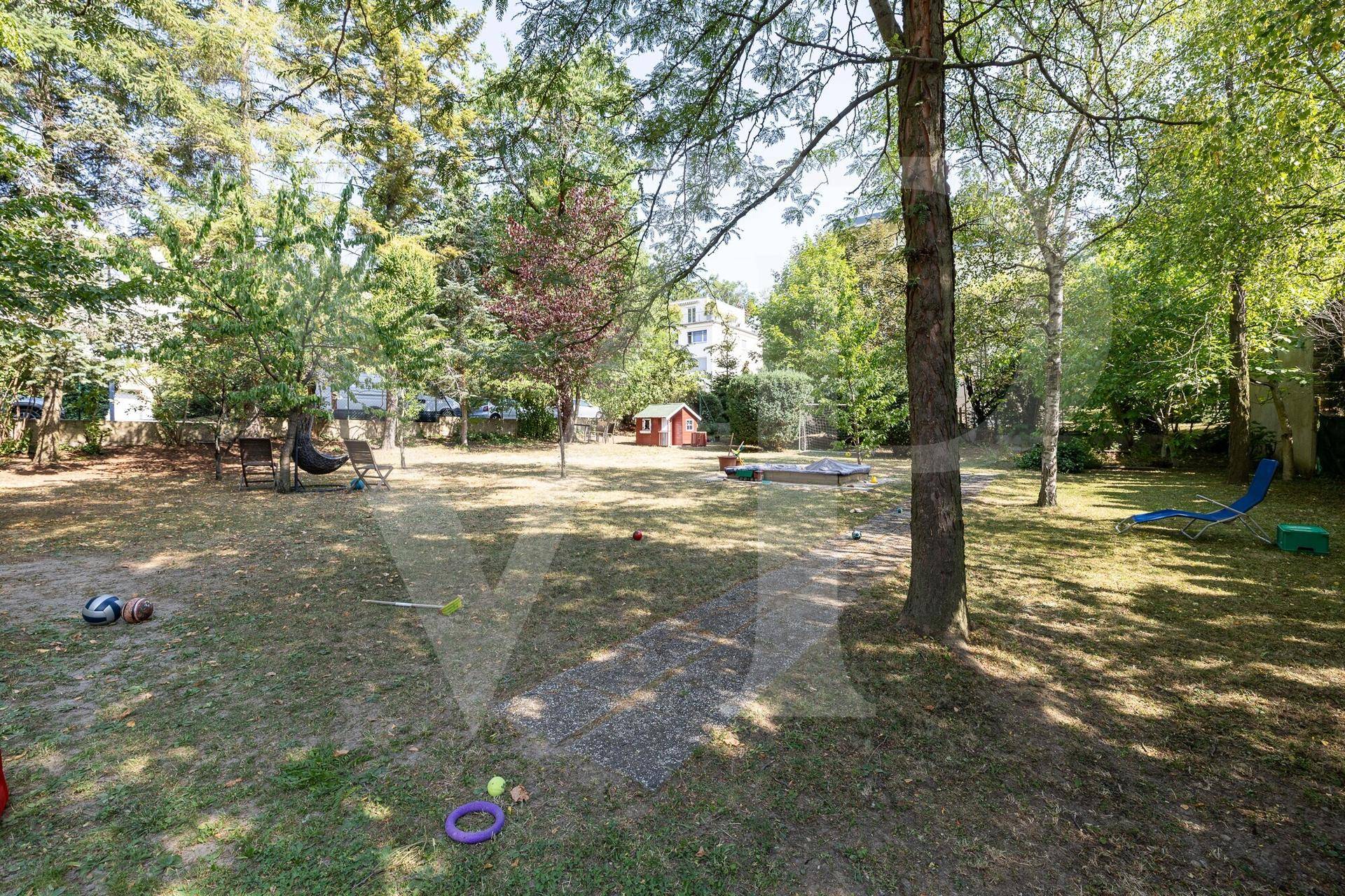 Großer, gepflegter Garten mit Rasenfläche, altem Baumbestand, Spielgeräten und einem roten Gartenhaus.