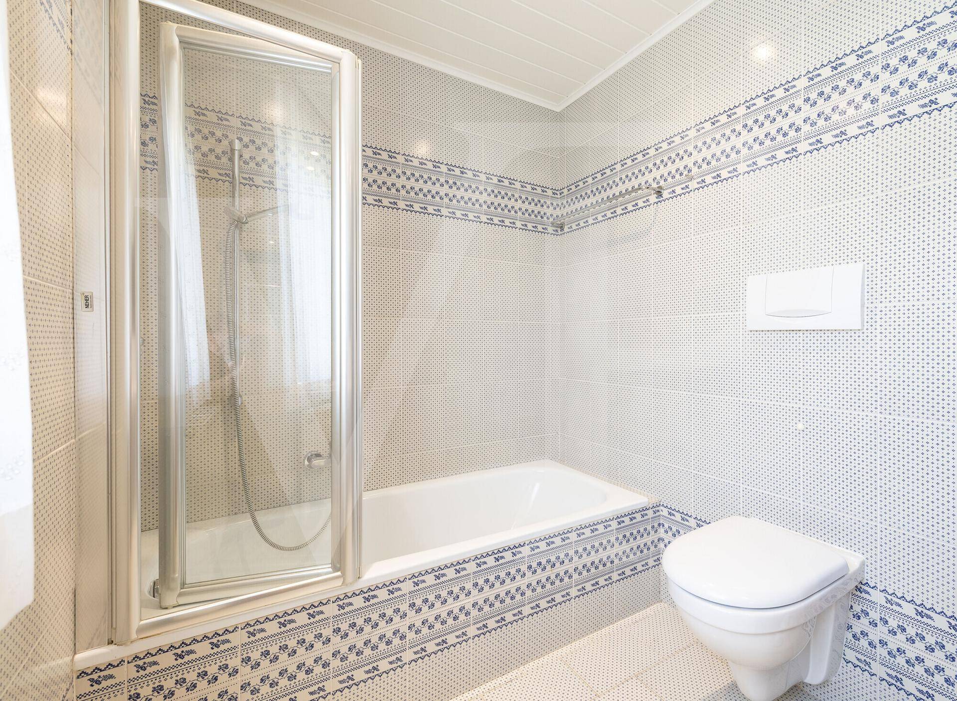 Funktionales Badezimmer mit Badewanne, Duschabtrennung und wandhängendem WC in blau-weißen Fliesen.