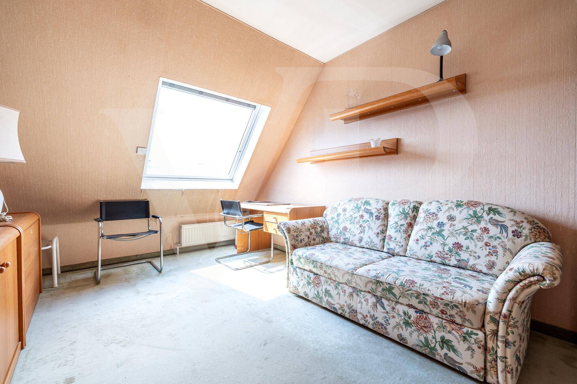Helles Dachzimmer mit Dachfenster, schräger Decke und gemütlichem Sofa mit Blumenmuster.