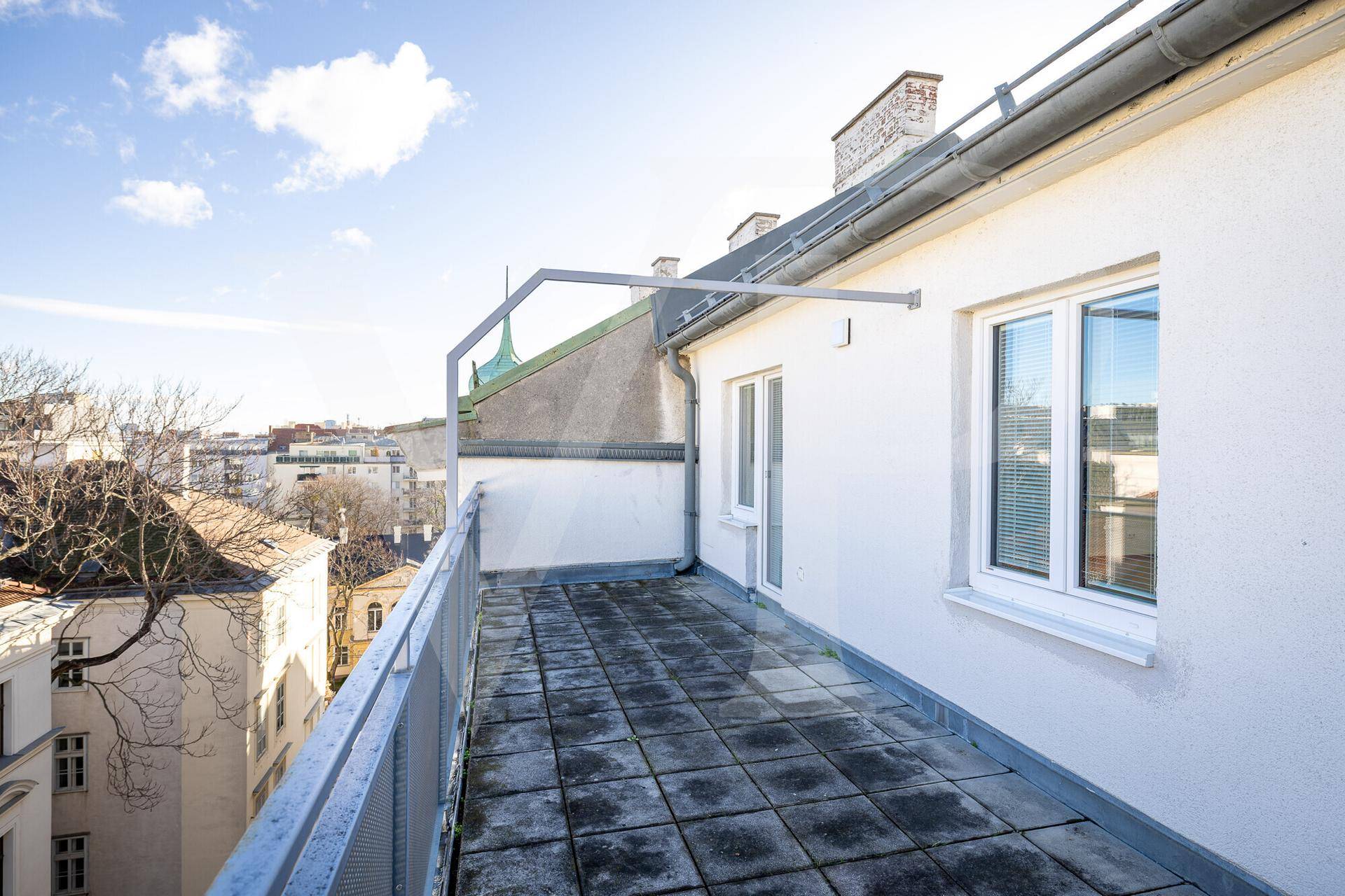 Schmale Terrasse mit Betonplatten und Metallgeländer, umgeben von städtischer Bebauung.