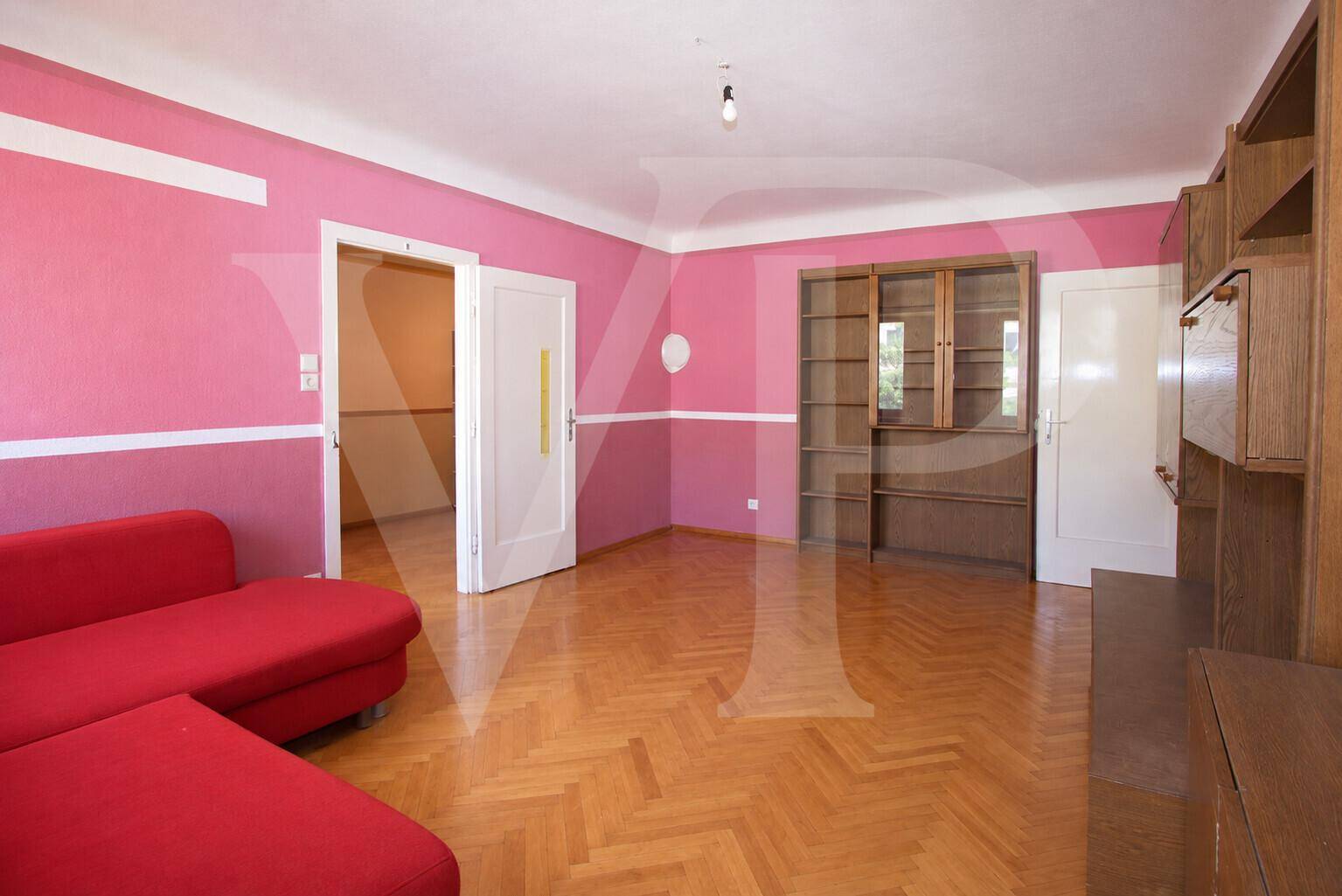 Lichtdurchflutetes Zimmer mit zwei großen Fenstern, weiß gestrichenen Wänden und Fischgrätparkett.