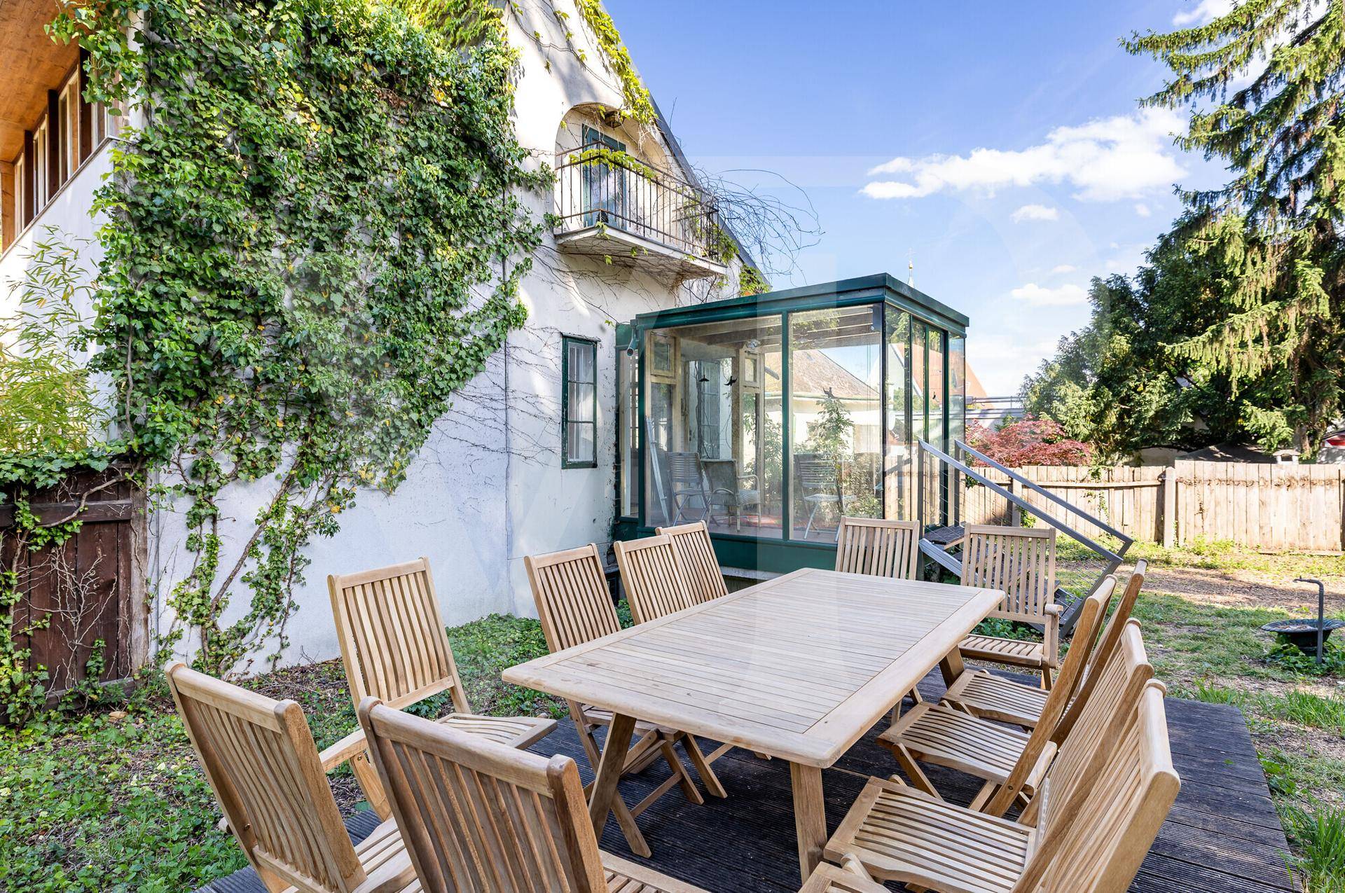 Gemütliche Holzterrasse mit Esstisch und Stühlen, angrenzend an einen Wintergarten und das Haus.