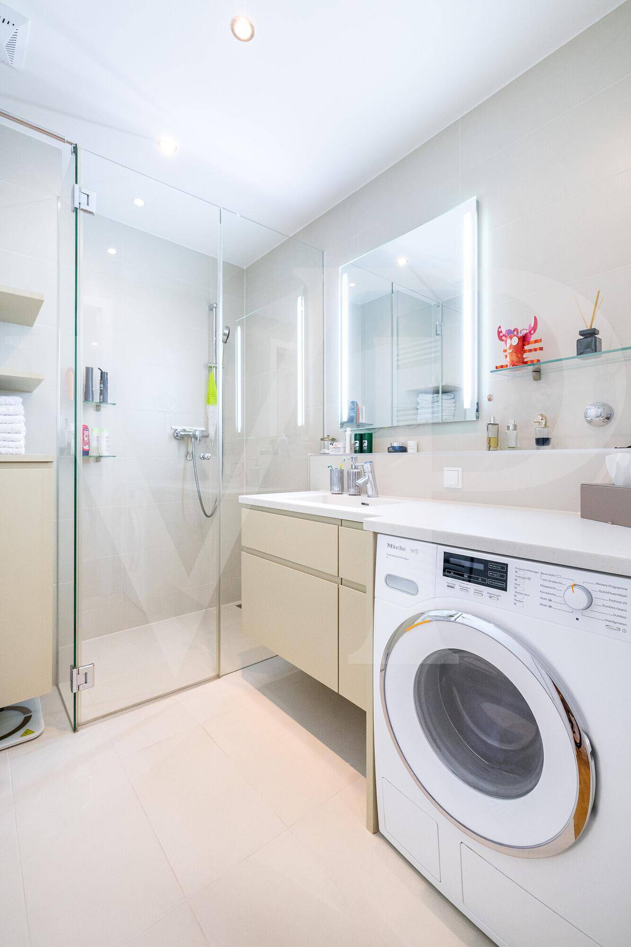 Modernes Badezimmer mit Dusche, Waschbecken und integrierter Waschmaschine, hell gefliest.