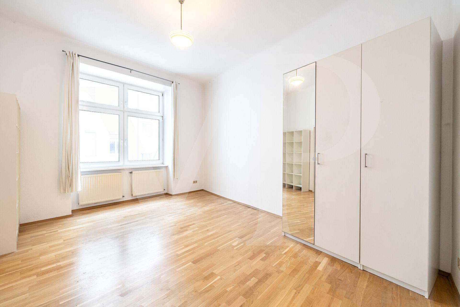 Geräumiges Zimmer mit hellem Parkettboden, großem Fenster und einem Spiegelschrank.