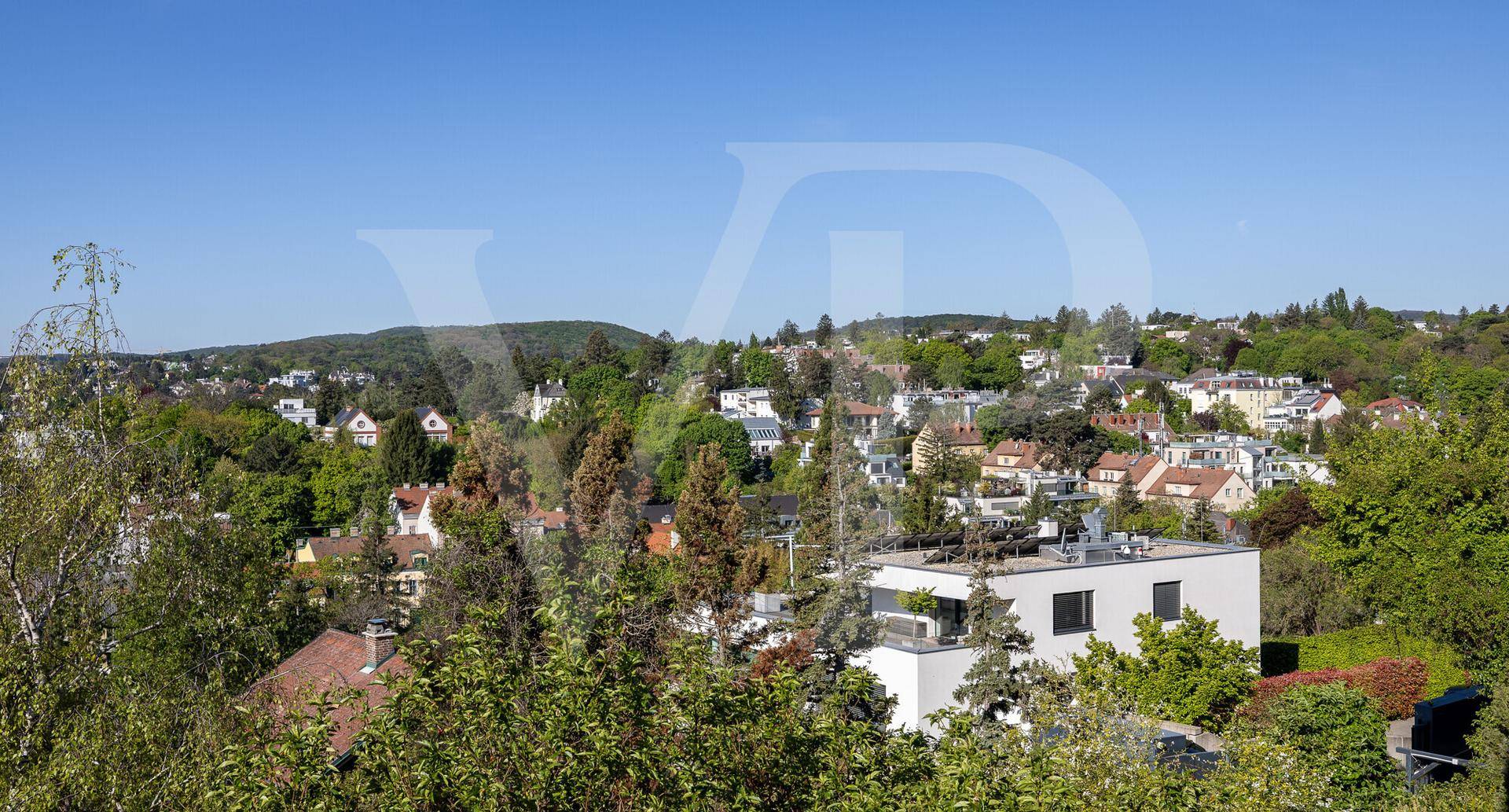 Blick über eine grüne Wohngegend mit einem modernen Gebäude im Vordergrund und Hügeln im Hintergrund.