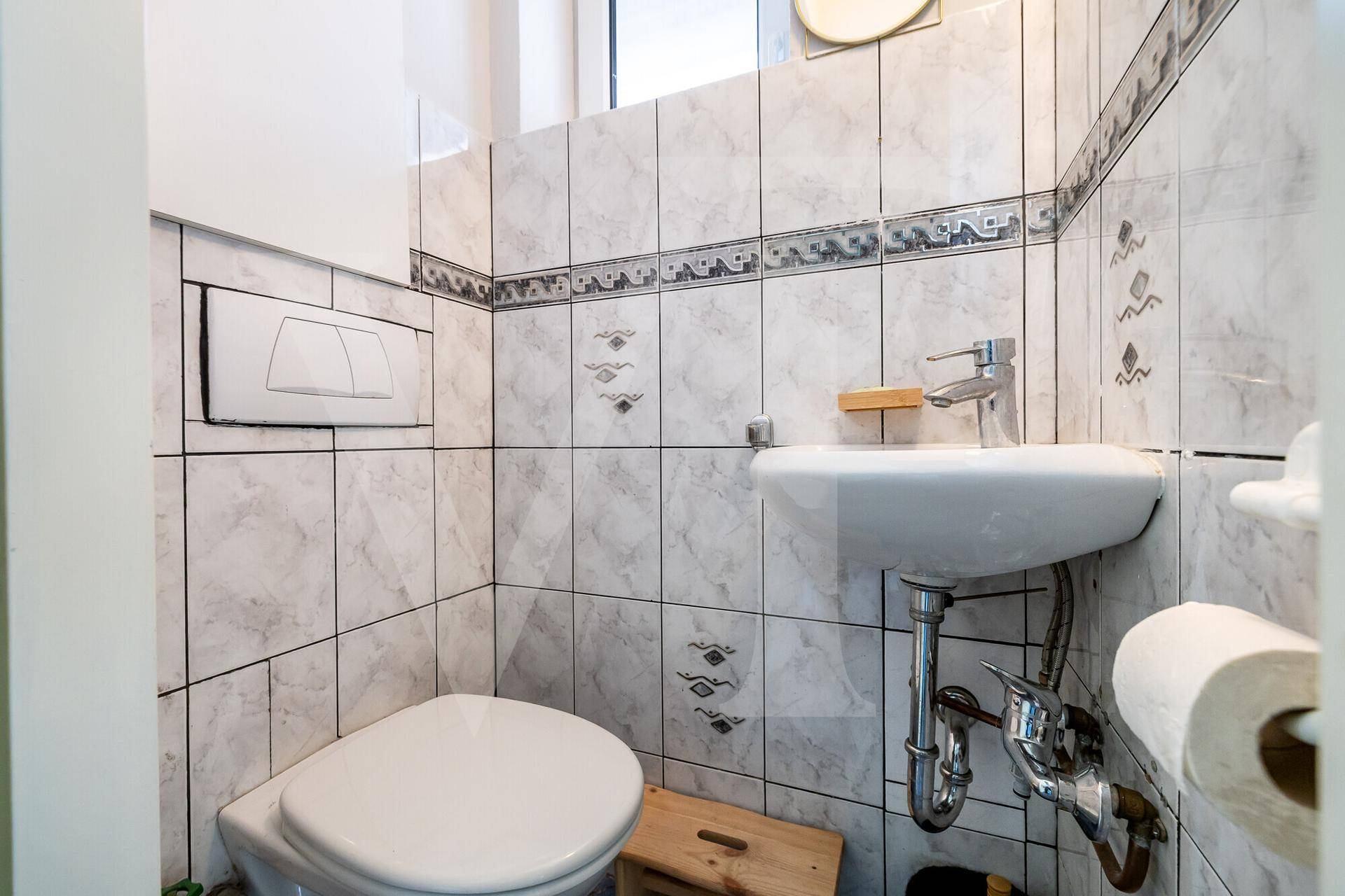Kompaktes Gäste-WC mit Toilette, kleinem Waschbecken und weiß-grauen Fliesen.