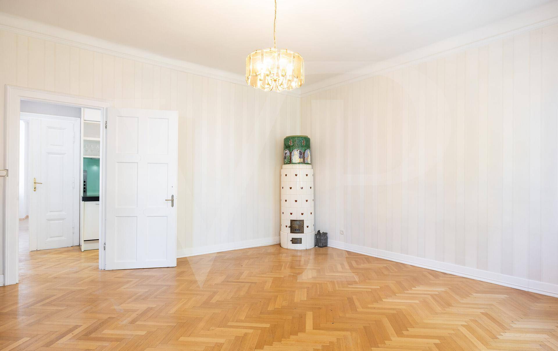Geräumiges Zimmer mit Fischgrätparkett, einem traditionellen Kachelofen und einem Kronleuchter.