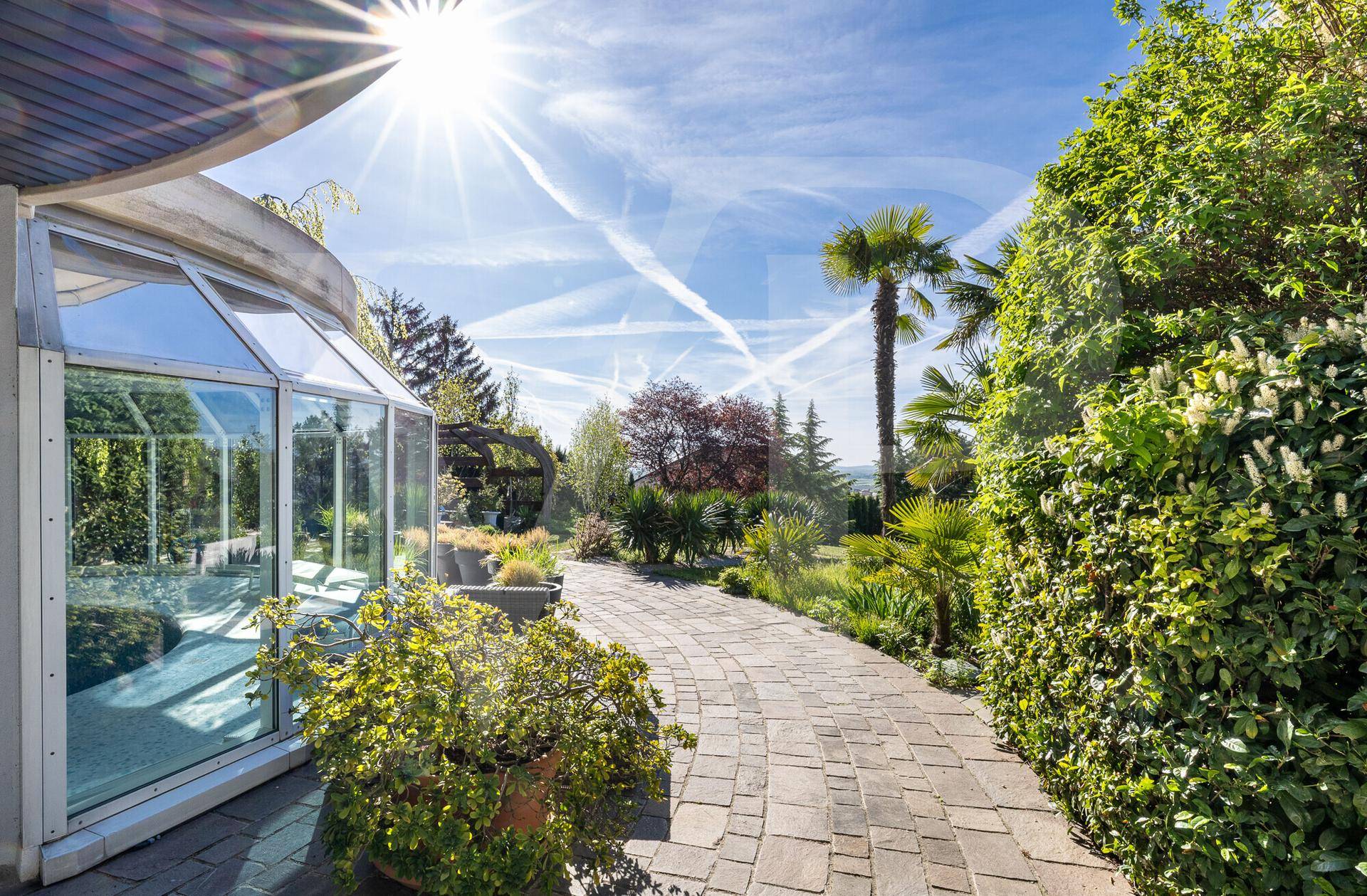 Sonniger Gartenweg mit exotischen Pflanzen, Palmen und einem Wintergarten.