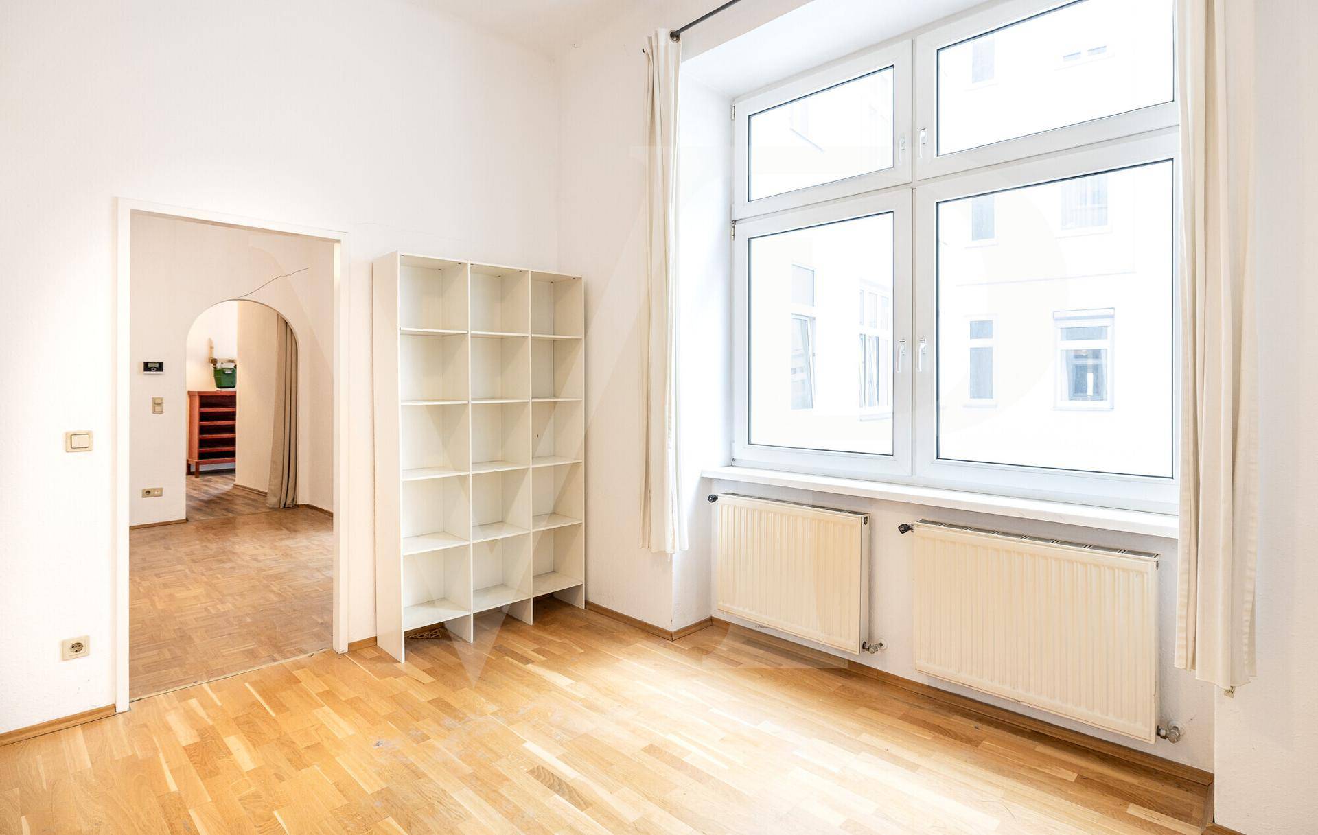 Heller Raum mit Parkettboden, großem Fenster und einem offenen Durchgang zu einem weiteren Bereich.