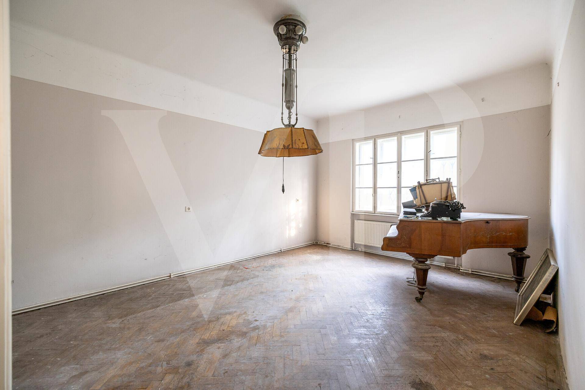 Zimmer mit abgenutztem Fischgrätparkett, hellem Wandanstrich, Fenster und einem Flügel.