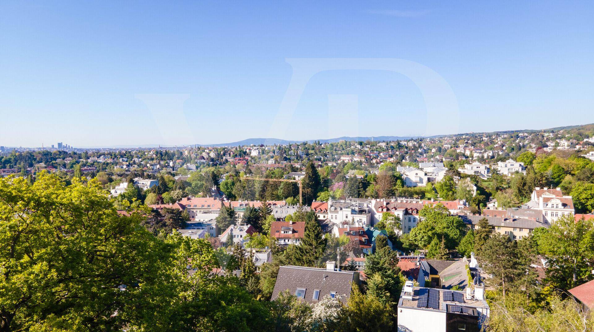 Weitläufige Ansicht einer grünen Wohngegend mit vielen Dächern und Bäumen unter blauem Himmel.