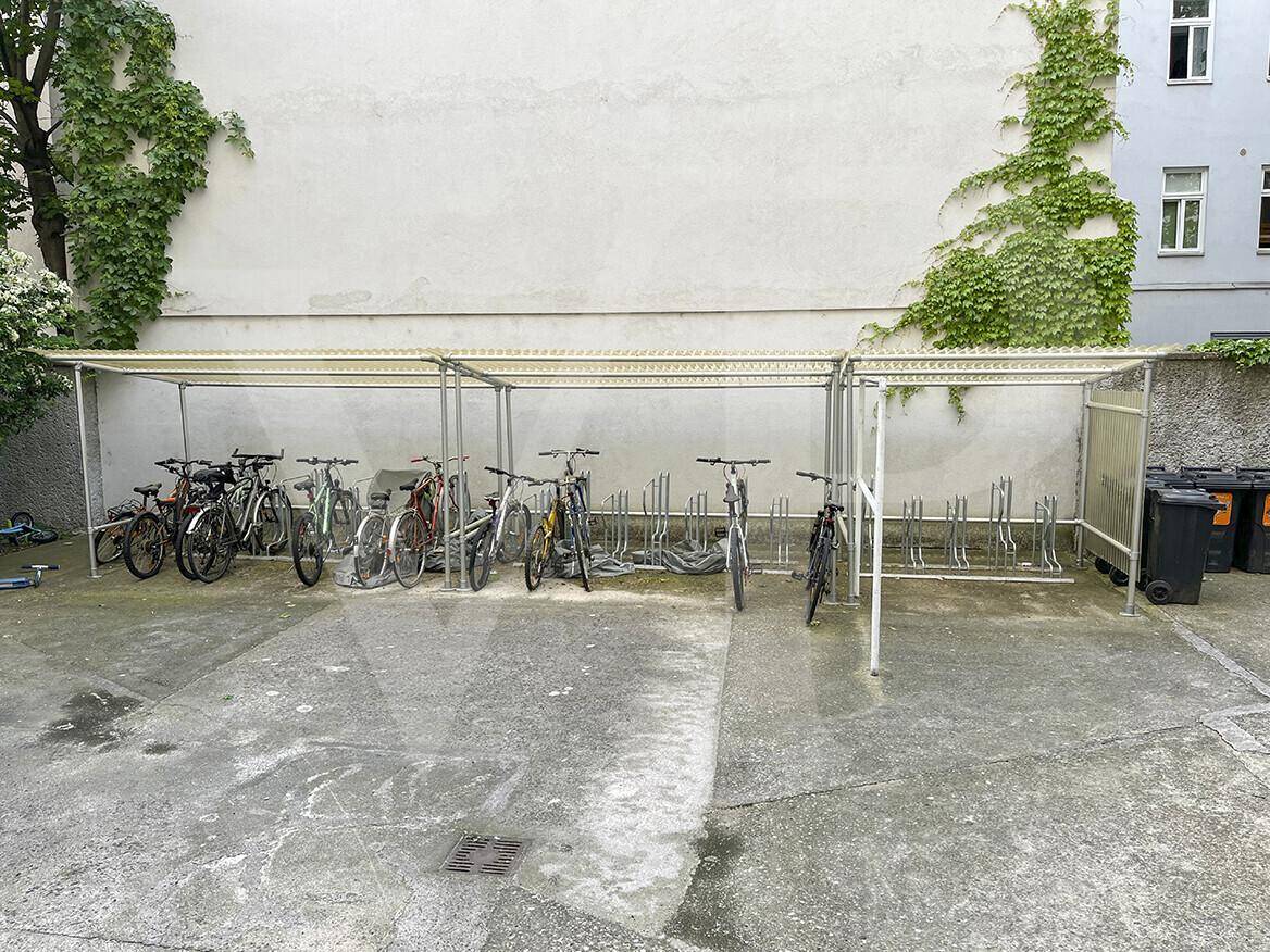 Überdachter Fahrradabstellplatz im Freien mit mehreren Fahrrädern und Mülltonnen.
