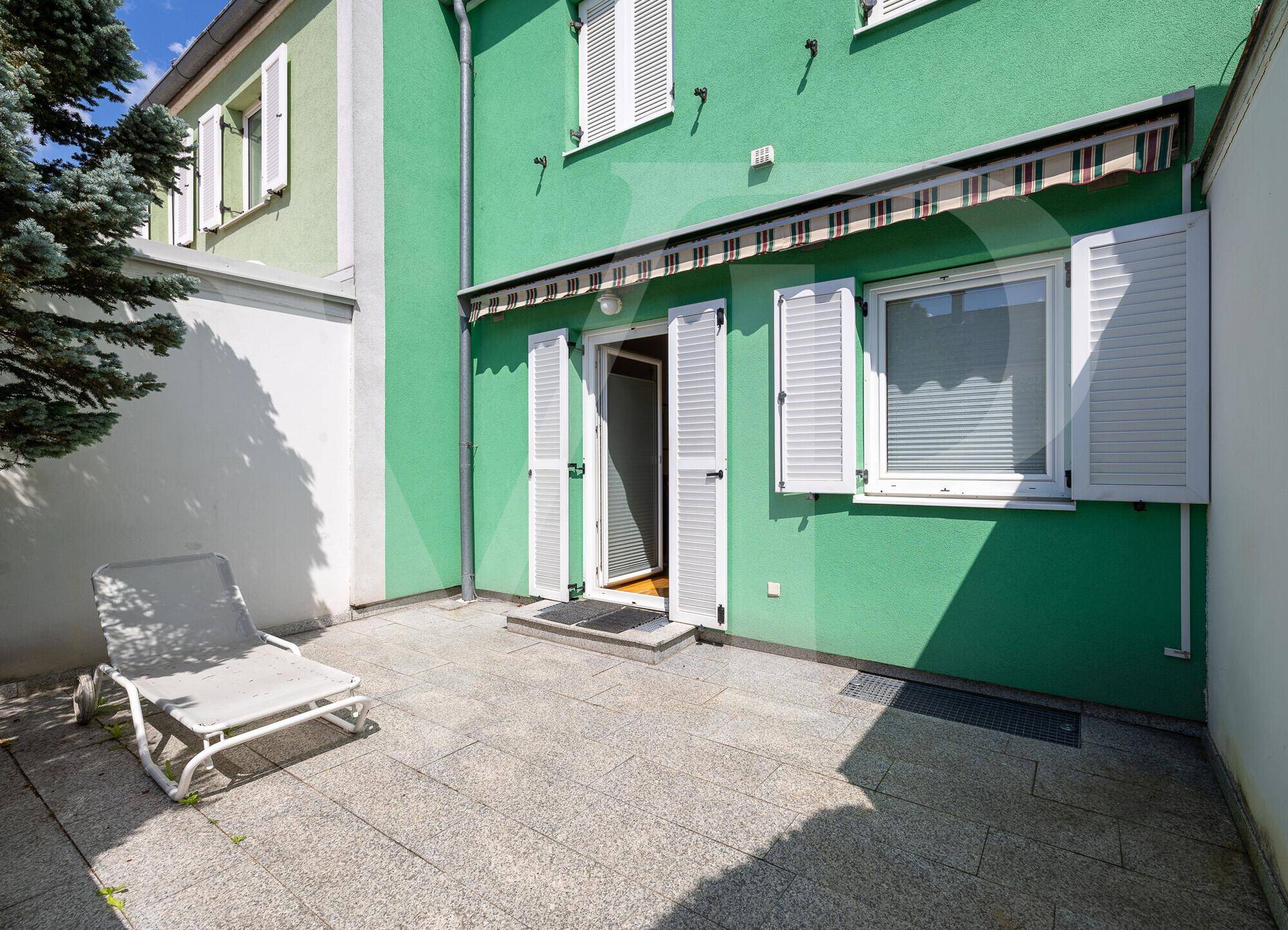 Sonnige Terrasse mit Steinplatten und Liegestuhl, direkt am grünen Reihenhaus gelegen.