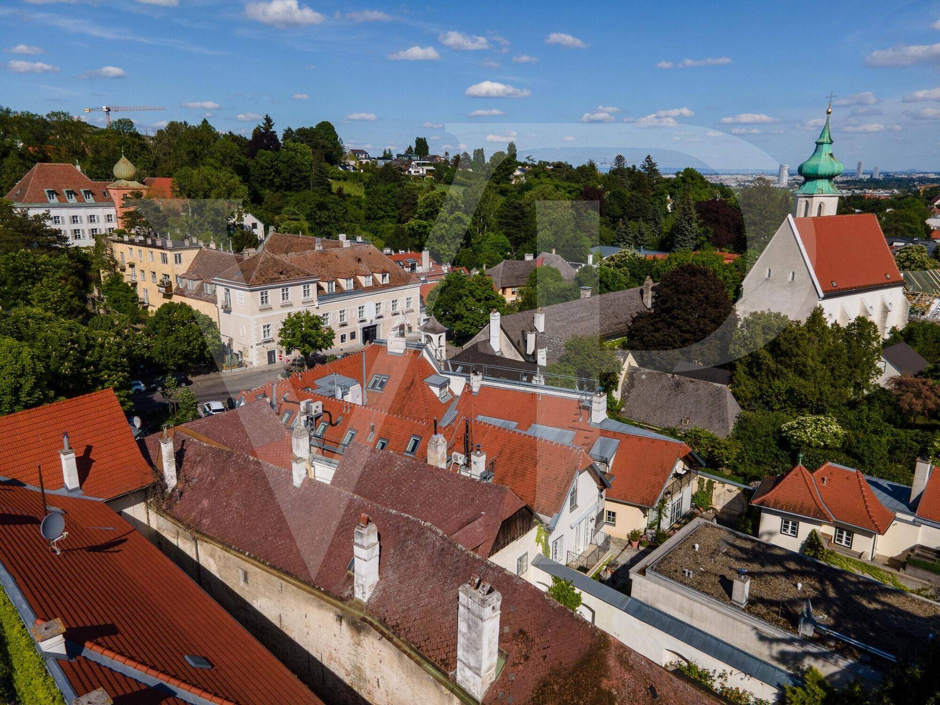 Malerische Stadtansicht mit roten Dächern, einer Kirche und grüner Landschaft unter blauem Himmel.