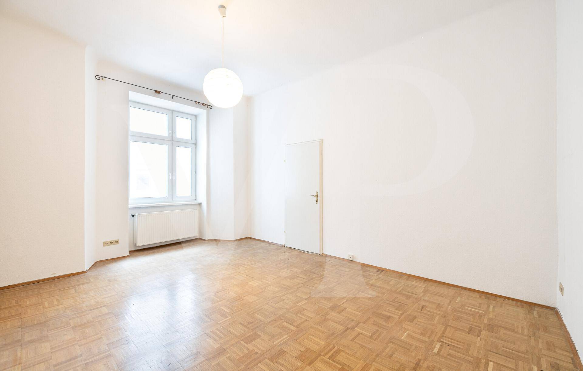 Geräumiges Zimmer mit Parkettboden, großem Fenster und einer weißen Eingangstür.